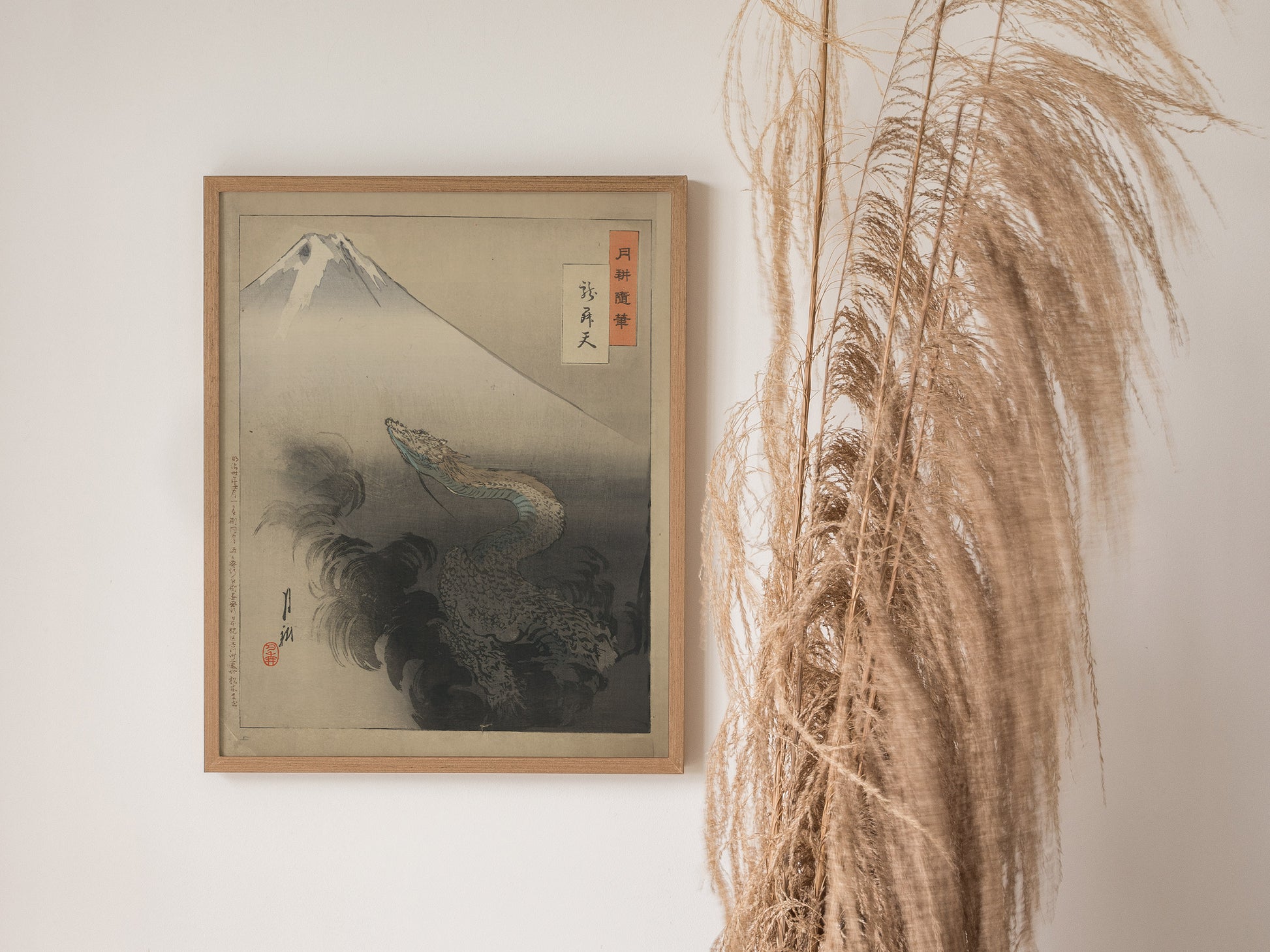 A framed Japanese art print features a dragon ascending through clouds with Mount Fuji in the background, displayed on a wall next to tall, dried ornamental grass.