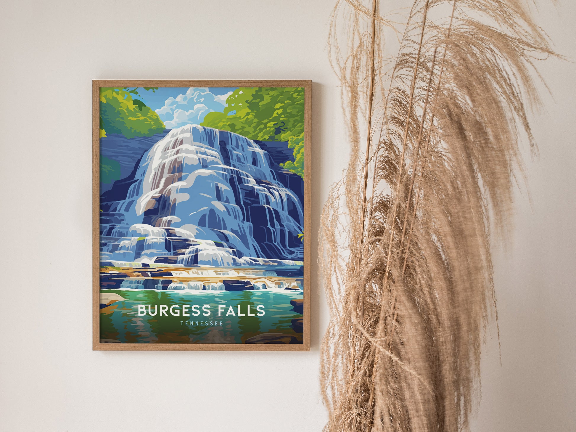 A framed poster of Burgess Falls in Tennessee hangs on a white wall, depicting a vibrant waterfall surrounded by greenery. Tall, dried grasses stand beside the artwork.
