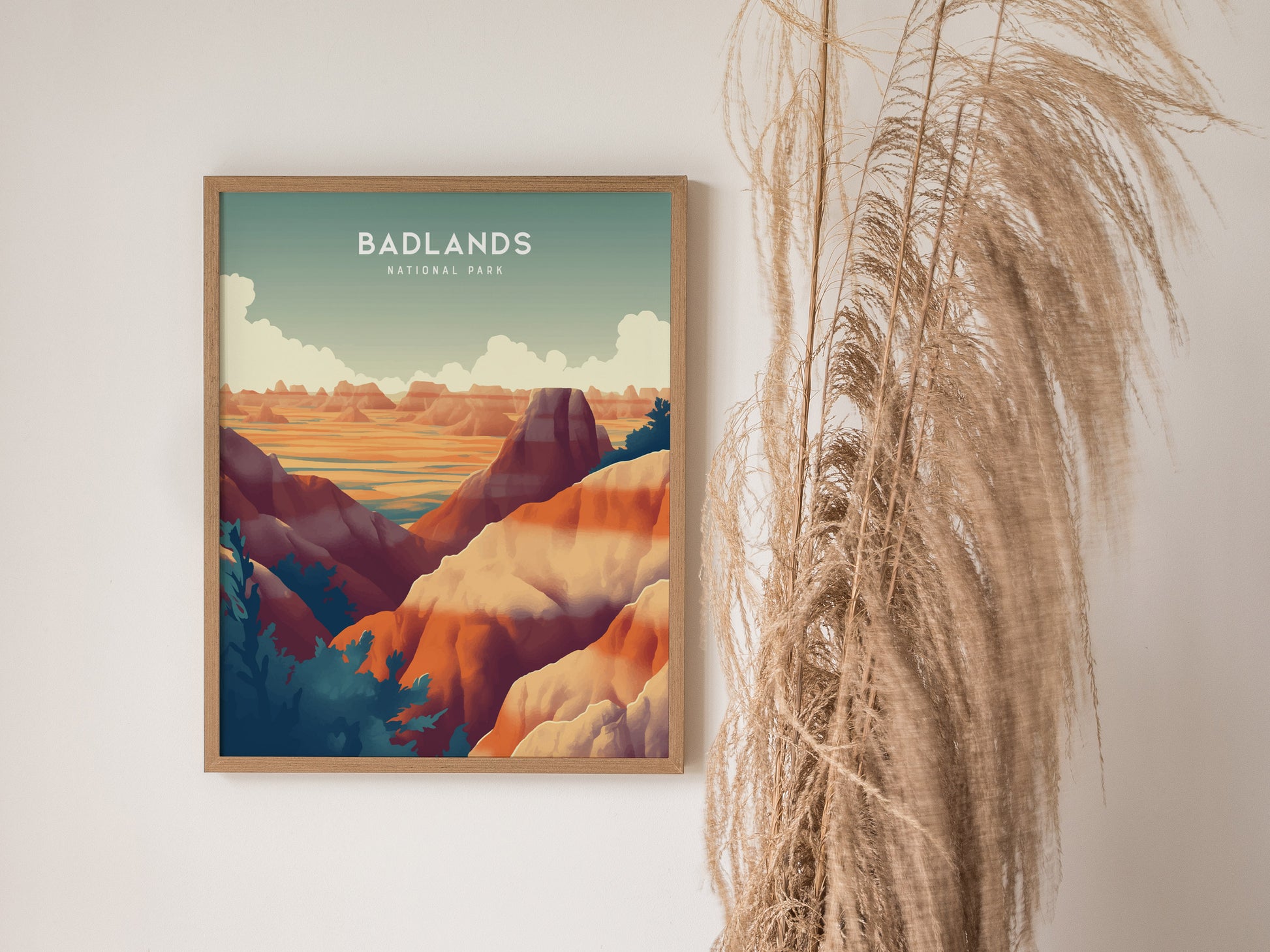A framed poster of Badlands National Park hangs on a white wall, depicting colorful rock formations. Tall, wispy dried grasses are positioned alongside the artwork.