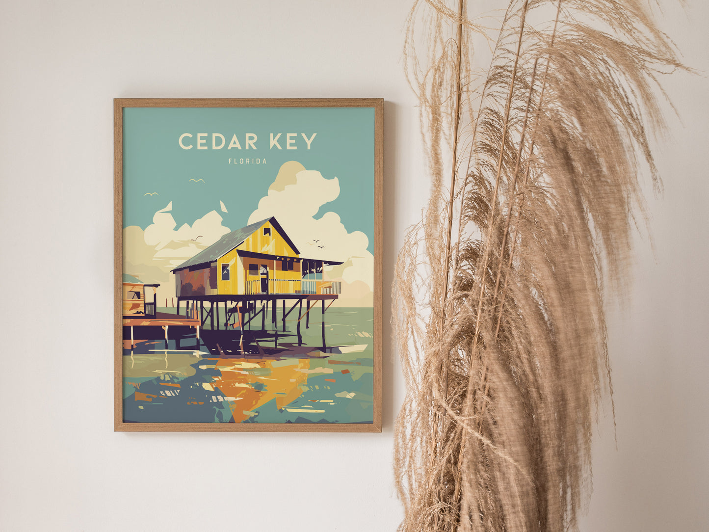 A framed Cedar Key, Florida poster depicting a stilt house above coastal waters hangs on a wall beside tall, dried pampas grass.