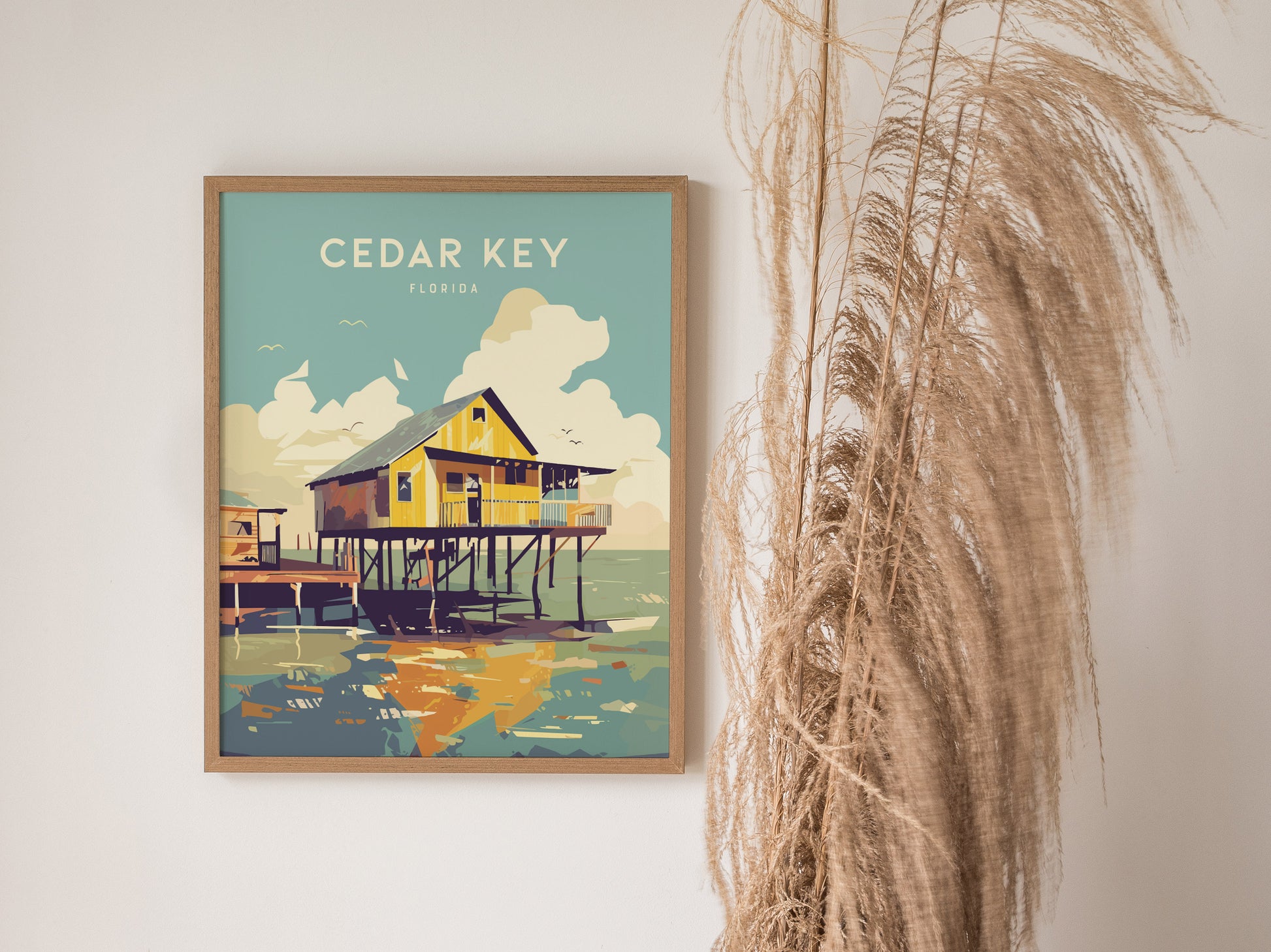 A framed Cedar Key, Florida poster depicting a stilt house above coastal waters hangs on a wall beside tall, dried pampas grass.