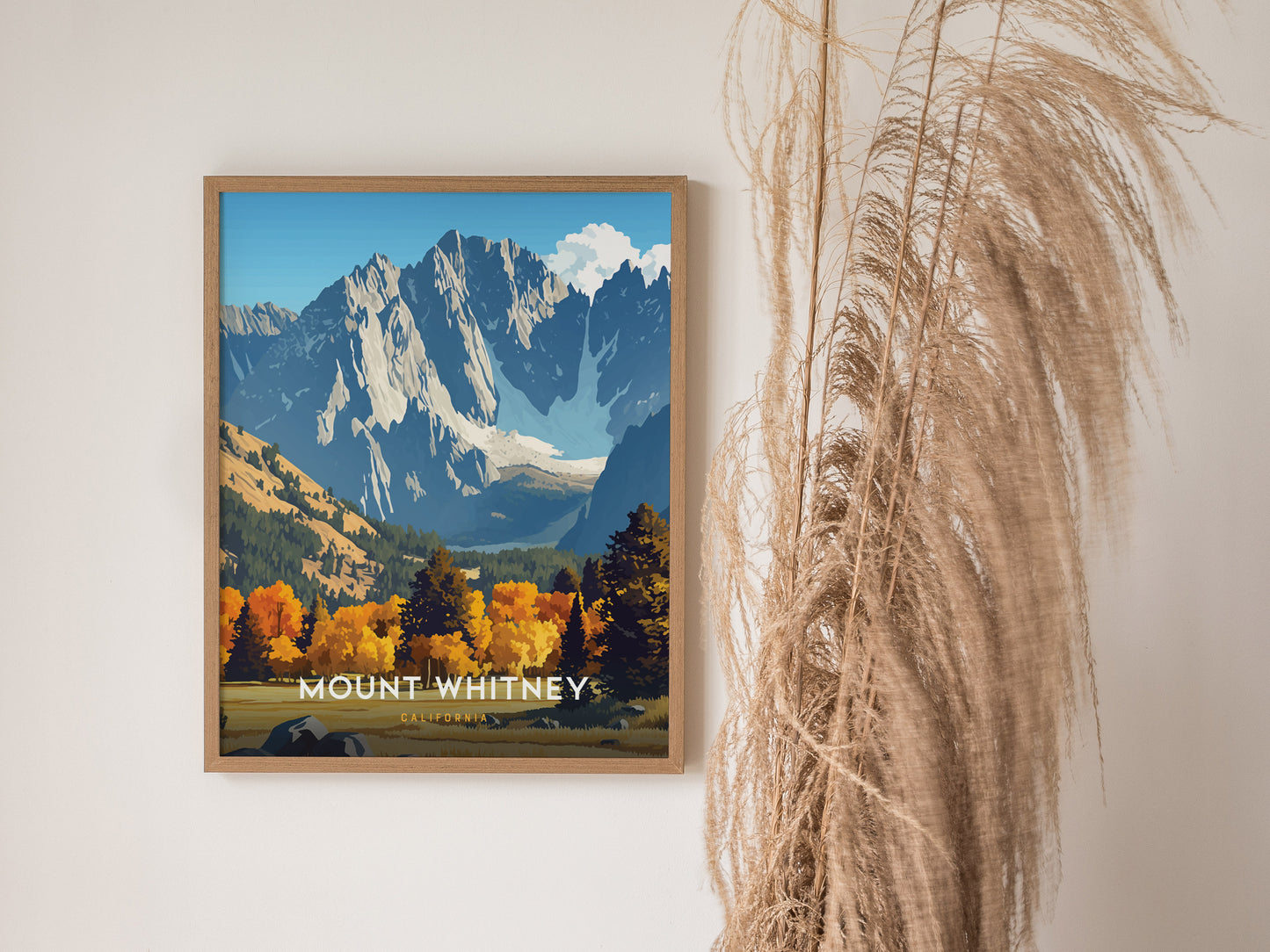 A framed poster of Mount Whitney featuring dramatic granite ridgelines and vibrant autumn foliage, set against a clear blue sky, hangs on a wall beside dry pampas grass.