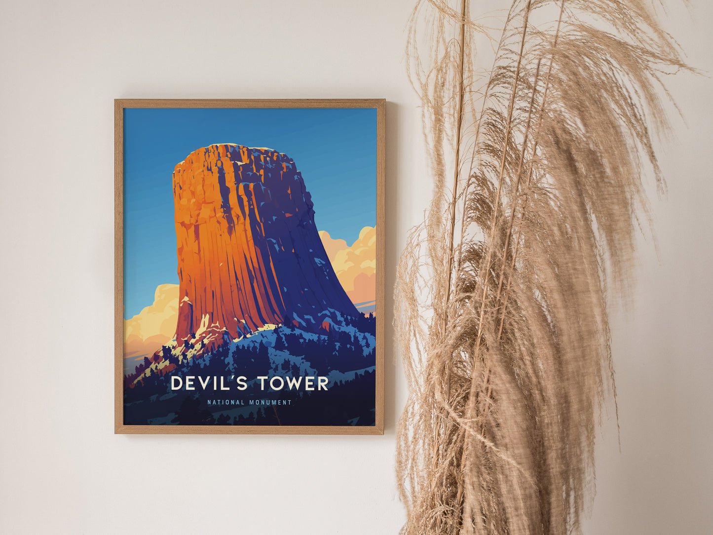 A framed poster of Devils Tower with vibrant colors is displayed on a wall beside tall, wispy pampas grass. The artwork captures the monument against a blue sky.