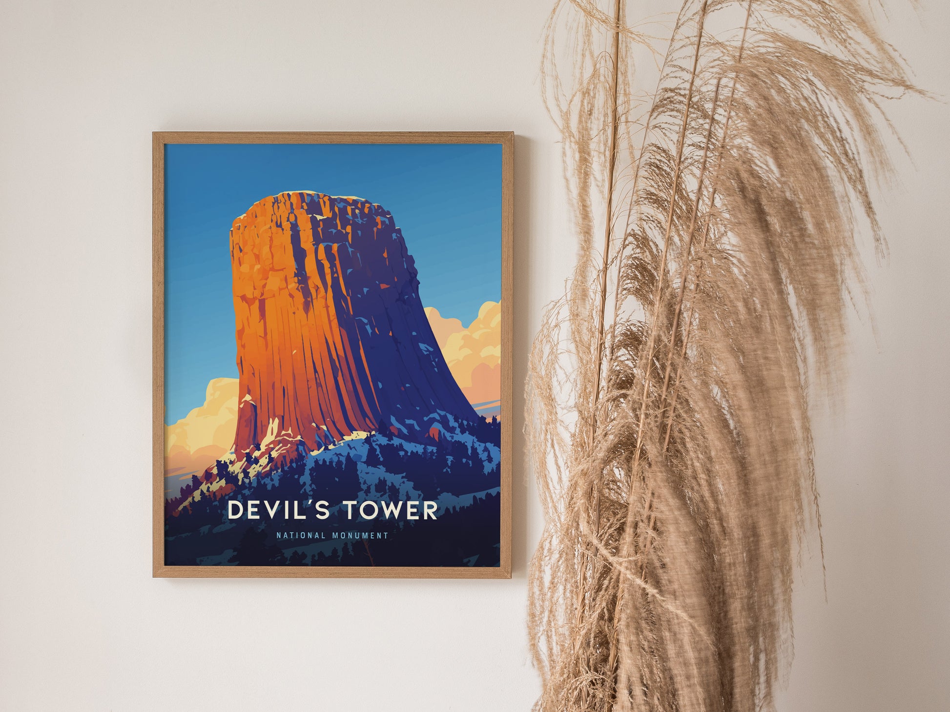 A framed poster of Devils Tower with vibrant colors is displayed on a wall beside tall, wispy pampas grass. The artwork captures the monument against a blue sky.