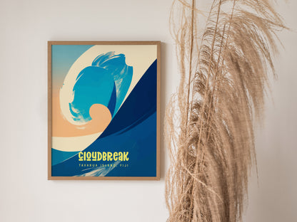 A framed surf poster with abstract wave art in blue and peach tones hangs on a white wall beside dried pampas grass. The text reads Cloudbreak, Tavarua Island, Fiji.