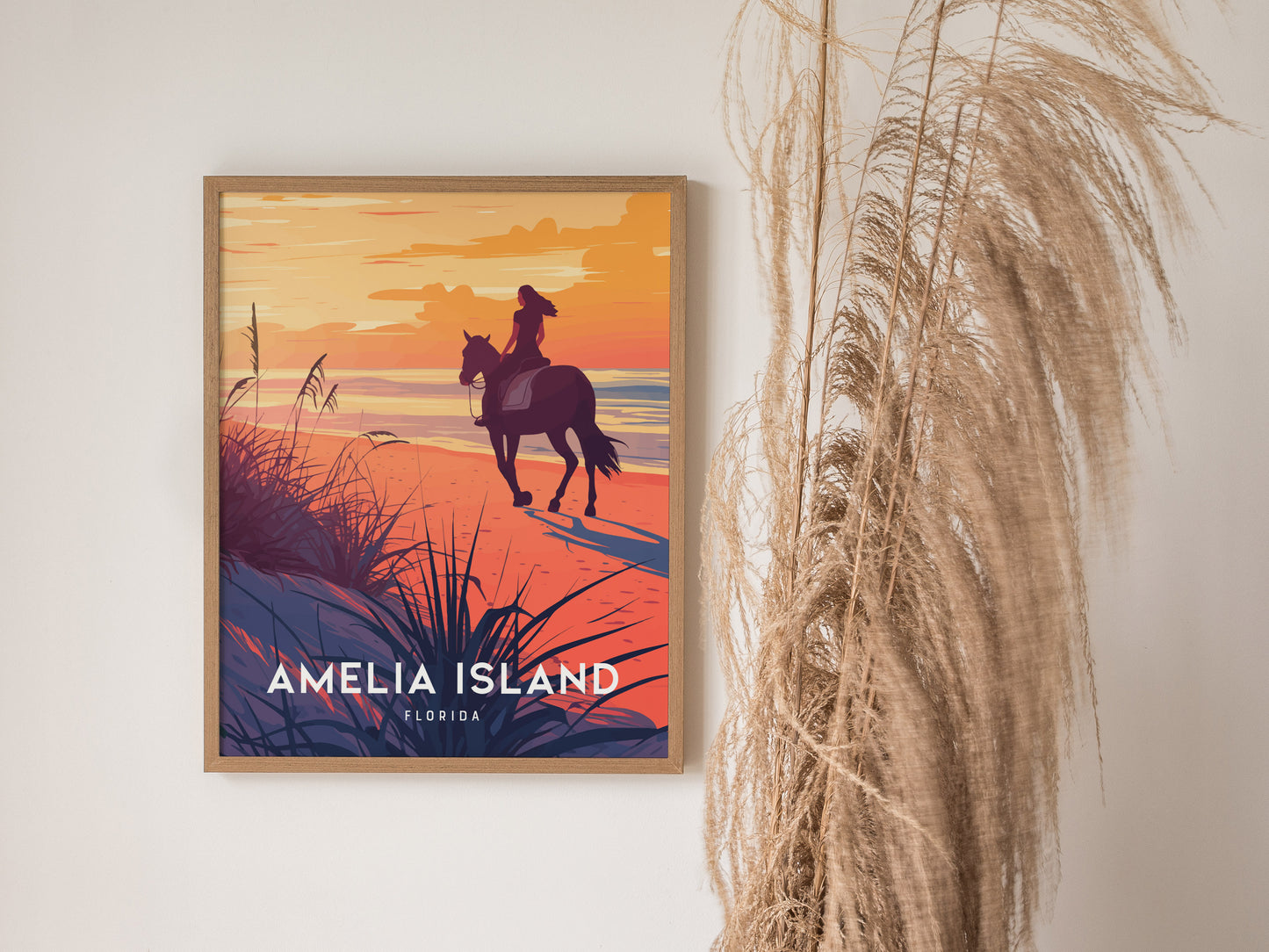 Framed Amelia Island poster depicting a woman on horseback at sunset, with vibrant orange and purple hues. Pampas grass decorates the wall beside the artwork.