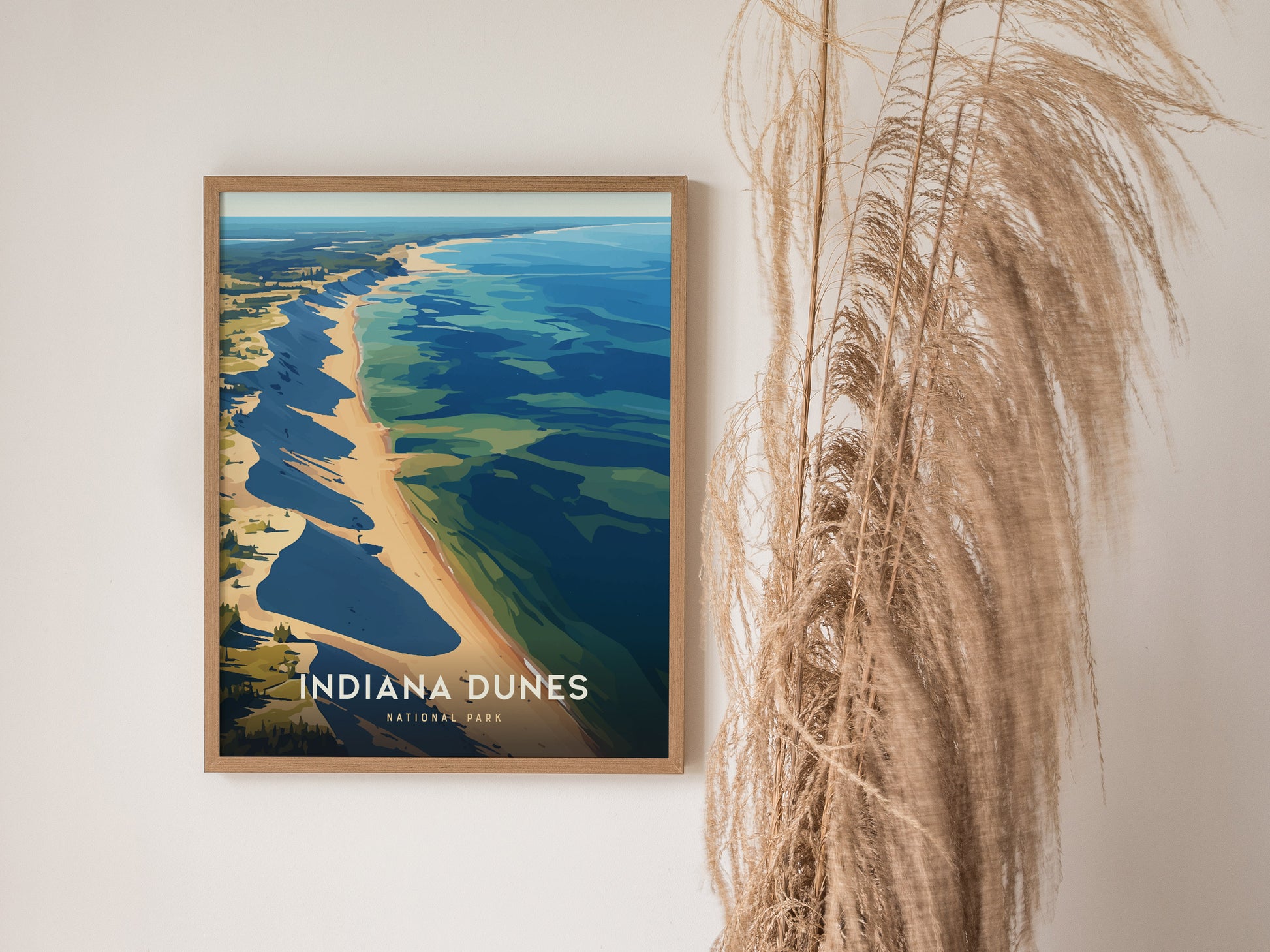 A framed poster of Indiana Dunes National Park hangs on a wall beside tall, dried pampas grass. The artwork depicts a scenic shoreline with vibrant blue.