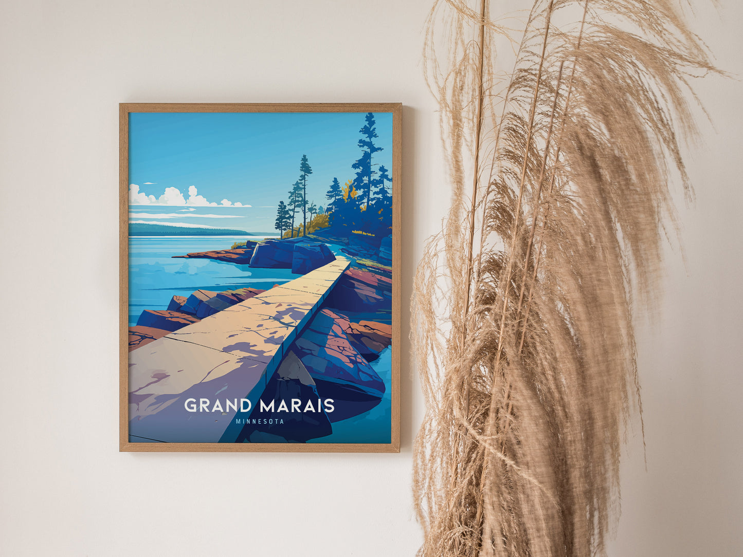 A framed poster of Grand Marais, Minnesota, featuring a vibrant landscape of a rocky shoreline and pine trees, hangs on a white wall next to tall dried grasses.