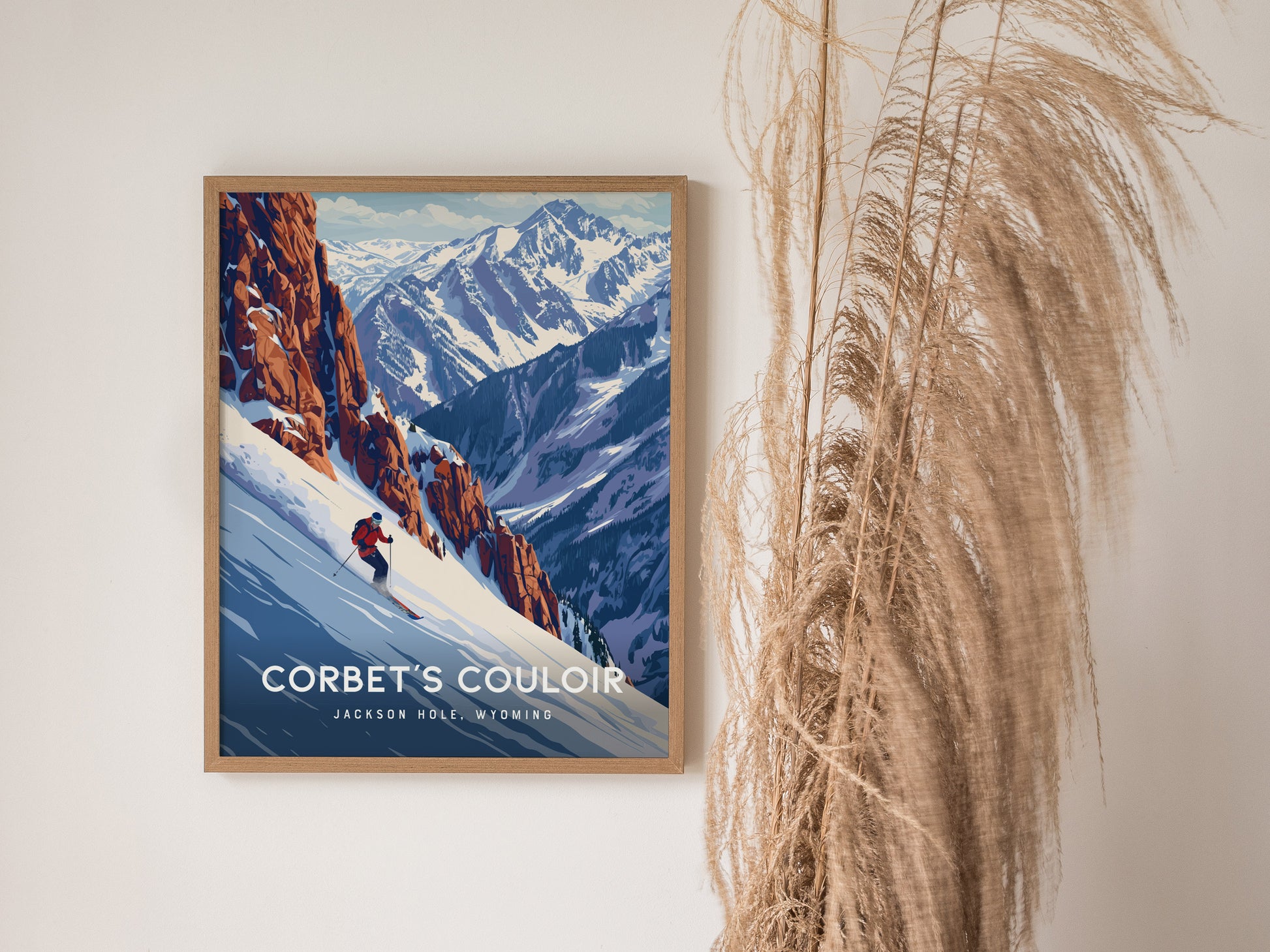 A framed poster of Corbets Couloir, featuring a skier descending a steep snowy slope with majestic mountain scenery, is displayed on a wall beside tall, dried pampas grass.