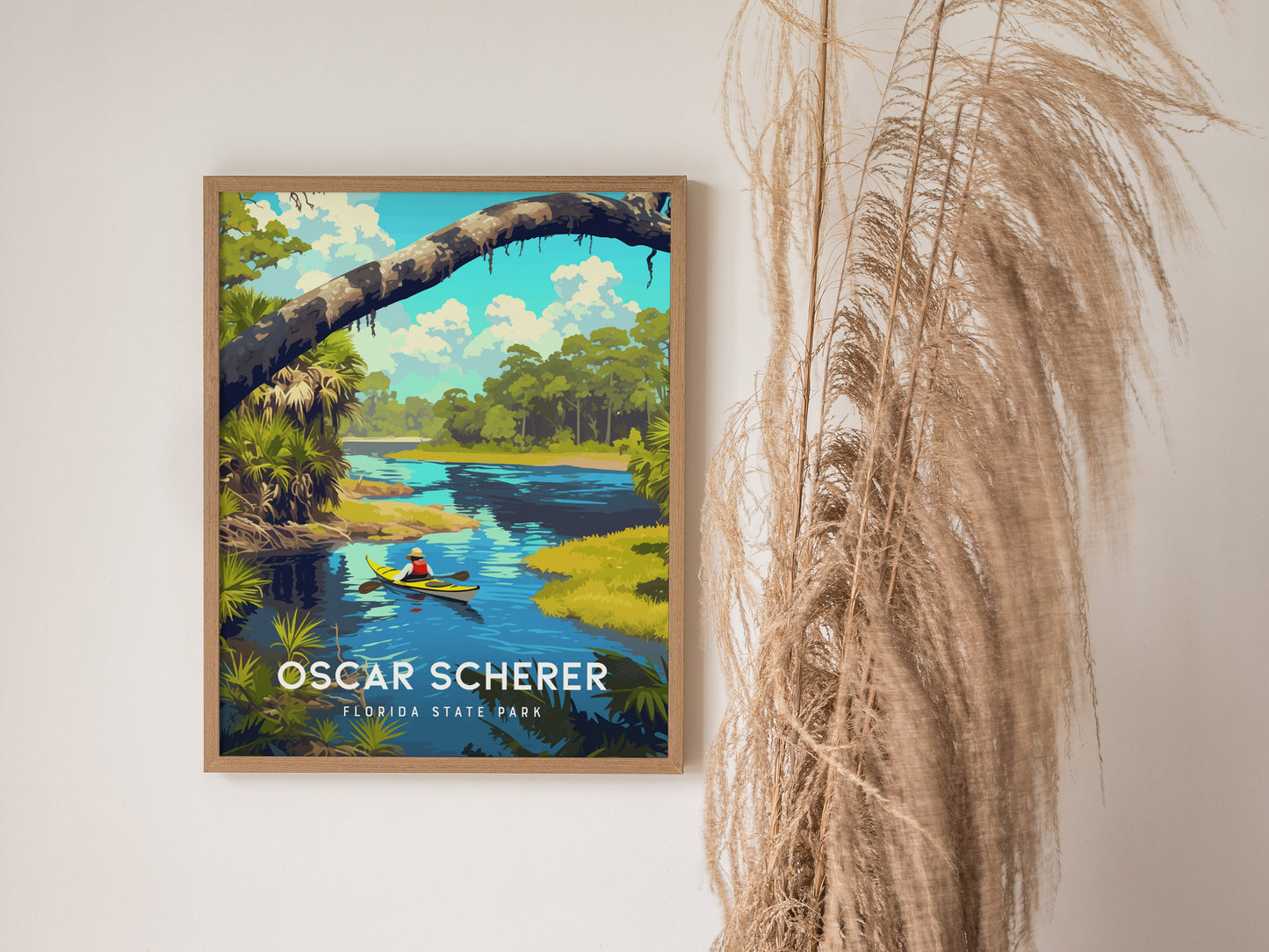 A framed poster of Oscar Scherer State Park features a vibrant illustration of a kayak on a tranquil river with lush greenery. Decorative grasses are placed beside the artwork.