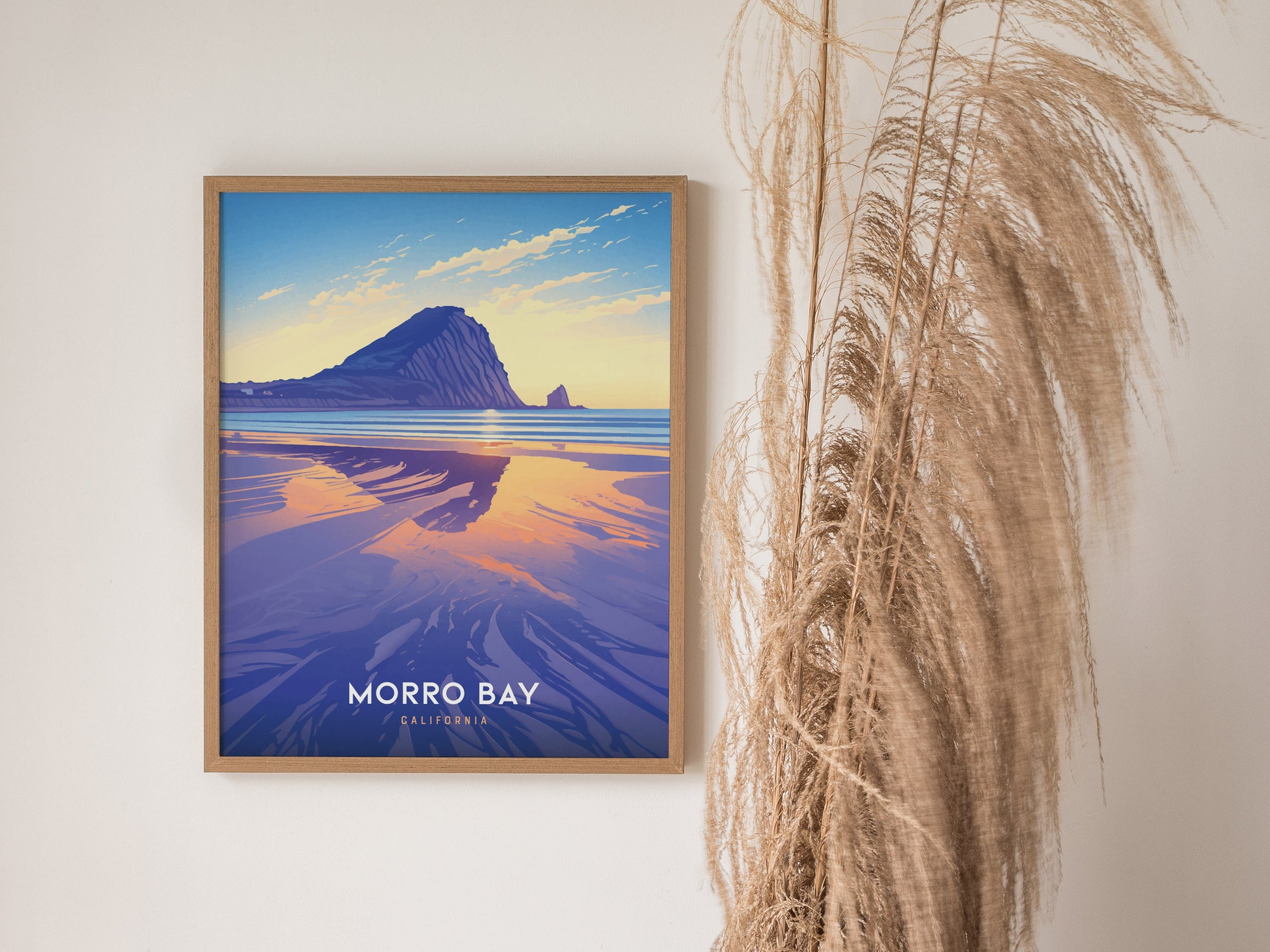 Framed poster of Morro Bay with a vibrant sunset reflecting on the water, featuring Morro Rock. Pampas grass decorates the wall beside the artwork, creating a serene atmosphere.