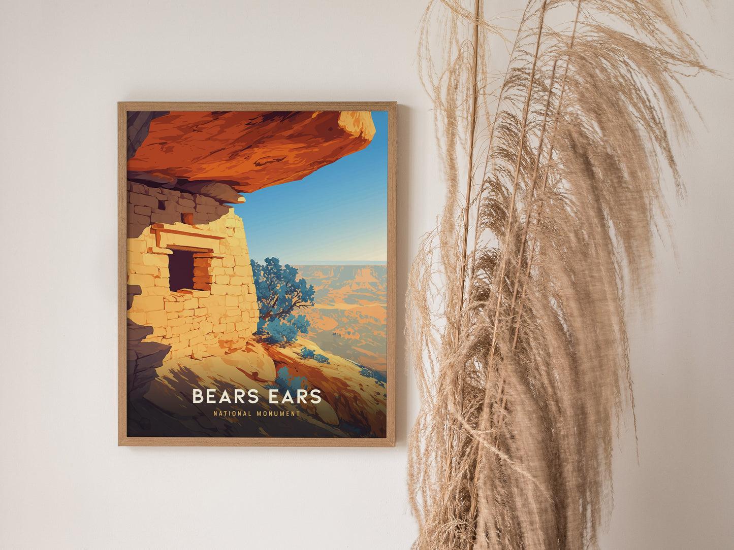 Framed poster of Bears Ears National Monument on a wall, featuring a vibrant depiction of red sandstone cliffs and ancient ruins. Dried pampas grass decorates the space beside it.