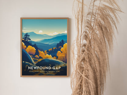 A framed poster of Newfound Gap in the Great Smoky Mountains features a colorful landscape with layered blue ridgelines and golden autumn trees. The image is set against a white wall with tall, wispy pampas grass nearby.