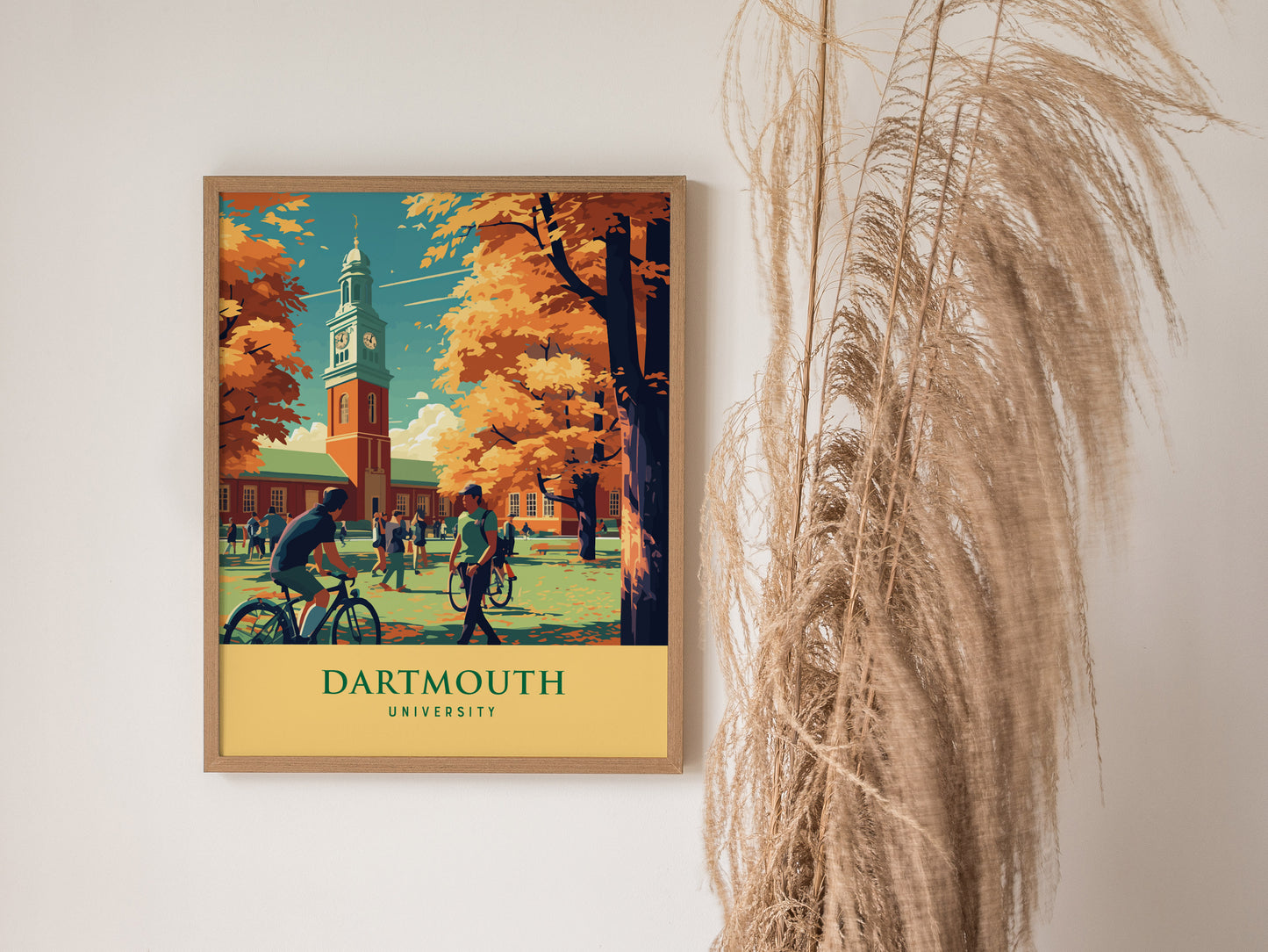 A framed Dartmouth University poster featuring a campus scene with students, bicycles, and autumn trees, hangs on a white wall beside dried pampas grass.