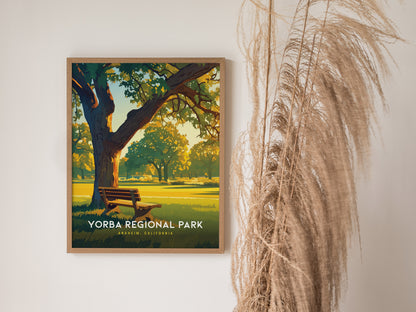 A framed Yorba Regional Park poster is displayed on a wall, showing a sunlit park scene with a bench and trees. Tall dried grasses are positioned beside it.