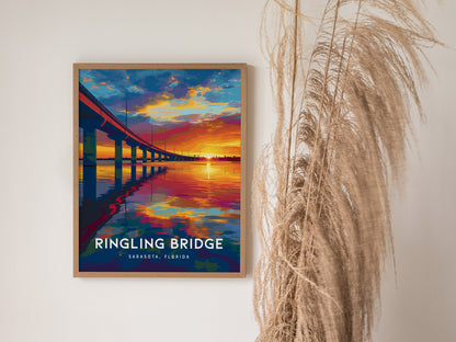 A colorful poster of the Ringling Bridge in Sarasota, Florida, is framed and hung on a white wall. The artwork depicts a vibrant sunset over the water, with the bridges reflection visible, accompanied by decorative dried grasses nearby.