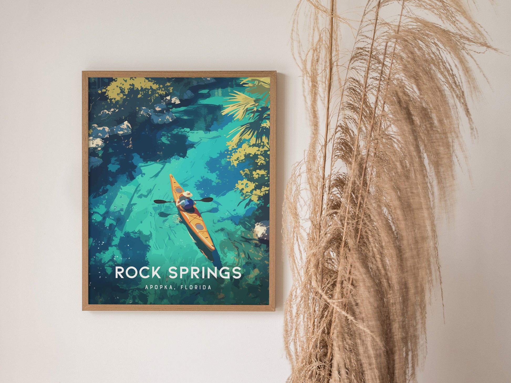A framed poster of a kayaker on turquoise waters, surrounded by lush greenery, with the text Rock Springs, Apopka, Florida. Pampas grass decorates the right side of the scene.