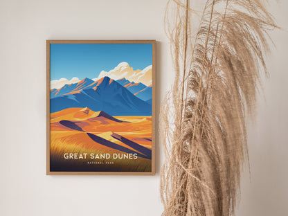 A framed poster of Great Sand Dunes National Park features vibrant yellow dunes and blue mountains, hanging on a wall next to tall, wispy dried plants.