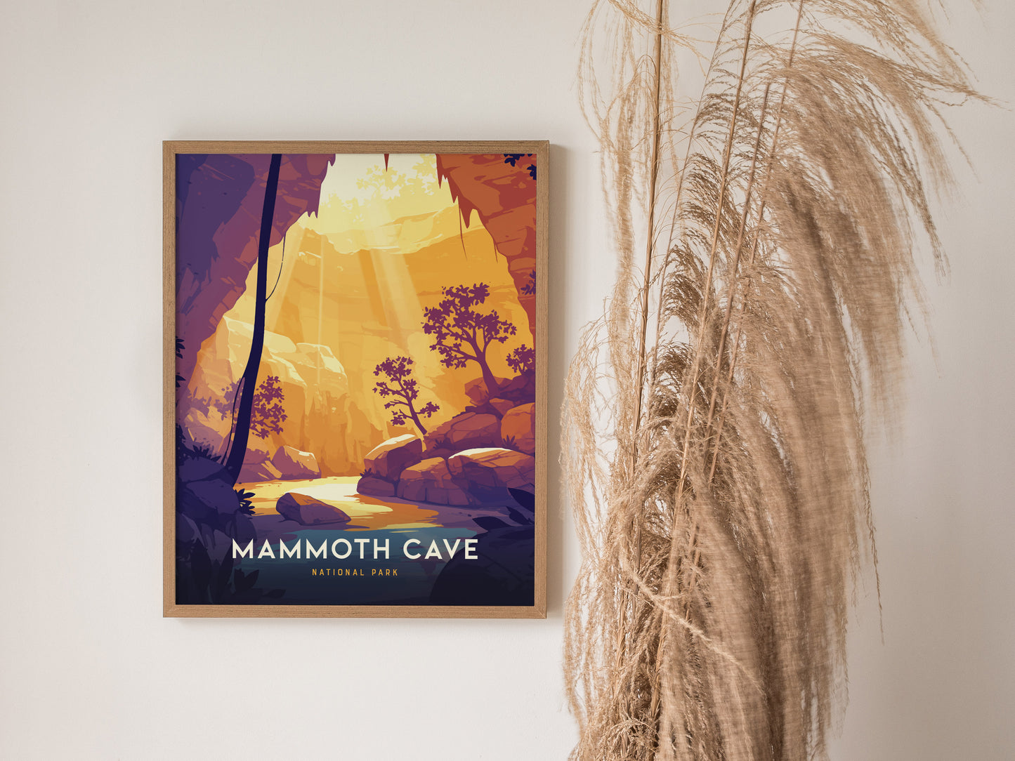 A framed poster of Mammoth Cave National Park features a stylized cave entrance with warm sunlight and silhouetted trees, hanging on a white wall beside tall, dried grasses.