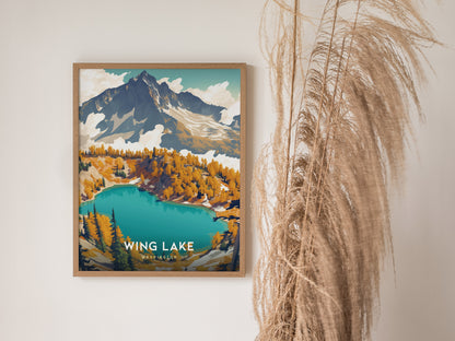 A framed poster of Wing Lake, Washington, depicting vibrant turquoise waters and autumnal trees with a mountain backdrop, is displayed beside tall, dried pampas grass.