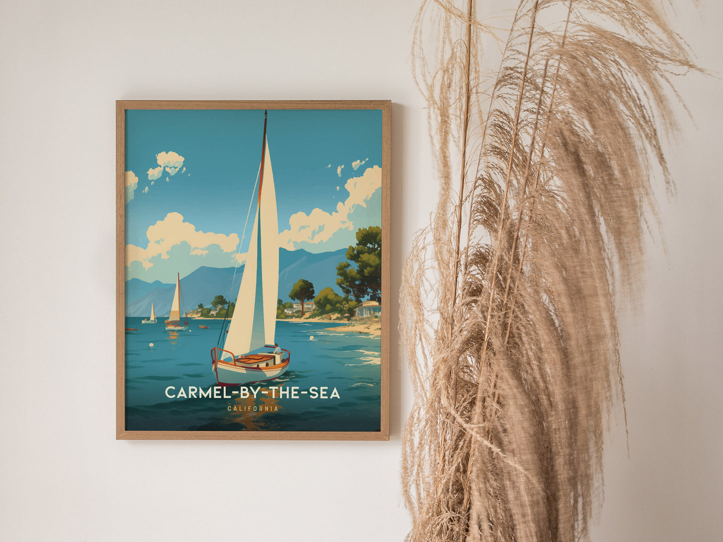 A framed poster of Carmel-by-the-Sea features a sailboat on a serene ocean, with mountains and trees in the background. Pampas grass decorates the right side of the image.