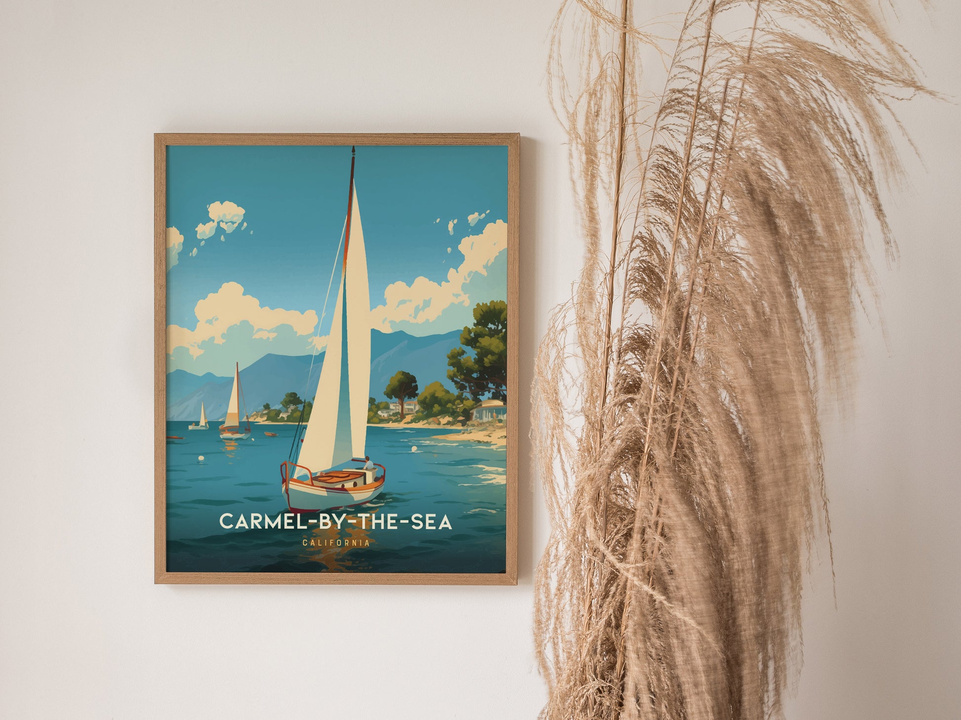 A framed poster of Carmel-by-the-Sea features a sailboat on a serene ocean, with mountains and trees in the background. Pampas grass decorates the right side of the image.