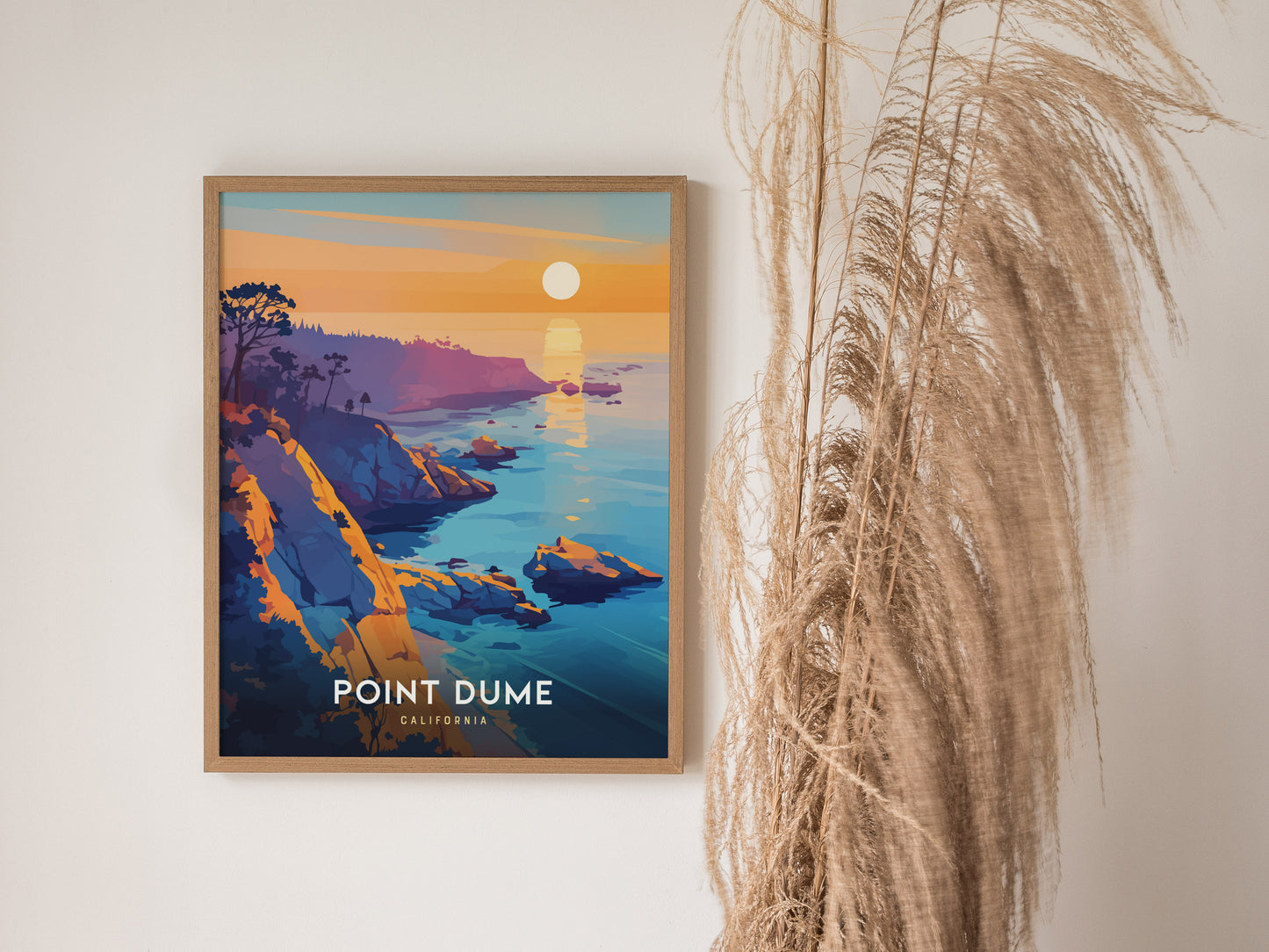 A framed poster of Point Dume, California, features vibrant cliffs and a sunset over the ocean, displayed next to pampas grass against a white wall.