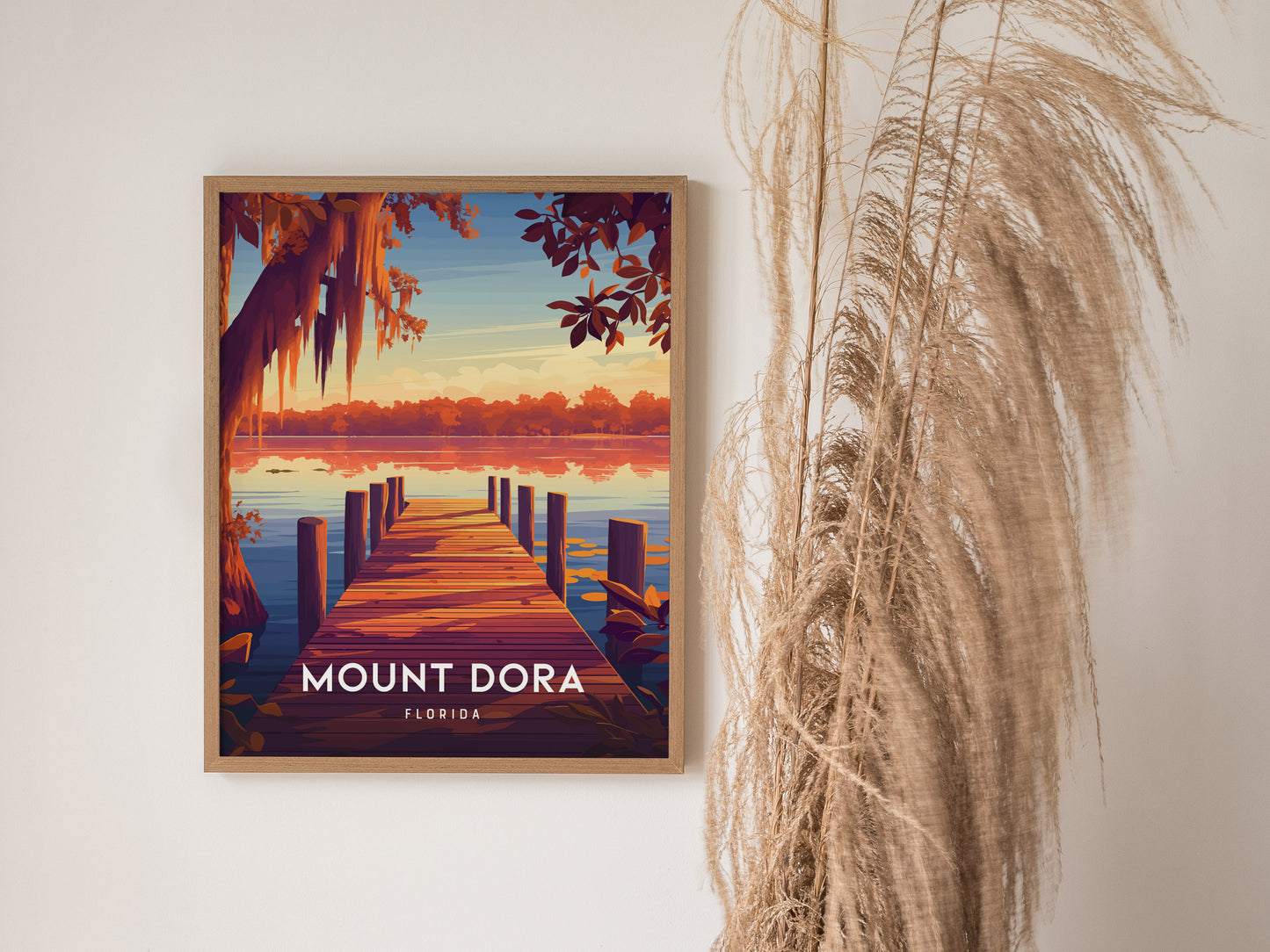 Framed Mount Dora poster depicting a serene lakeside view with a wooden dock and vibrant sunset colors. Decorative dried grasses are beside the artwork, enhancing a natural ambiance.