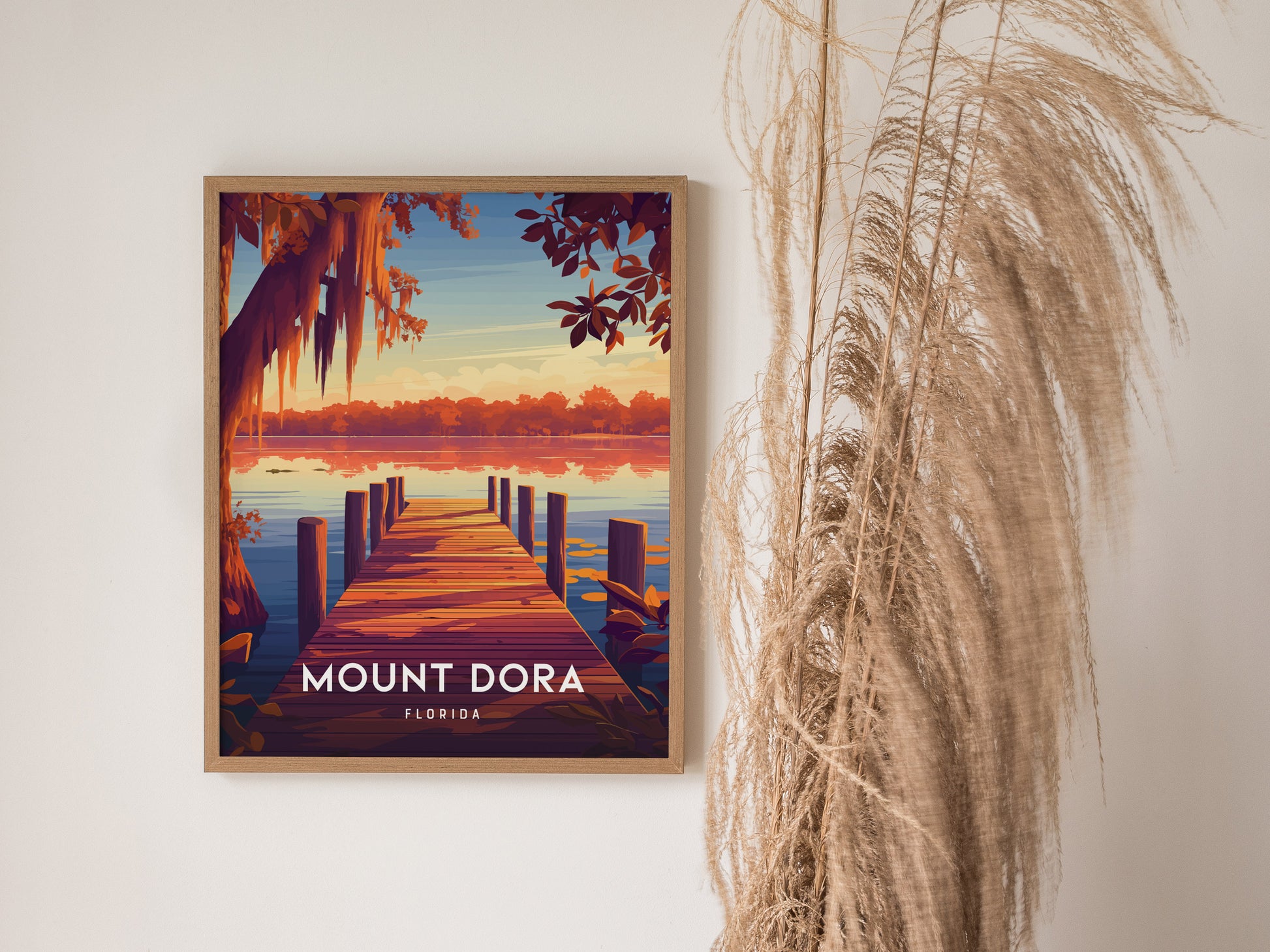 Framed Mount Dora poster depicting a serene lakeside view with a wooden dock and vibrant sunset colors. Decorative dried grasses are beside the artwork, enhancing a natural ambiance.