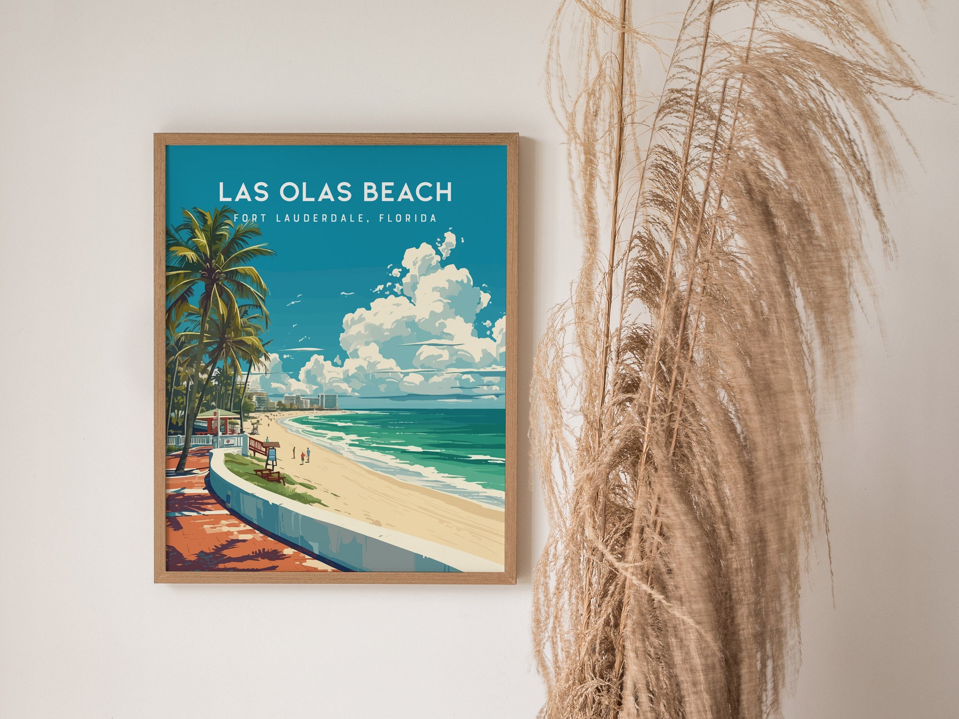 A framed poster of Las Olas Beach in Fort Lauderdale, Florida, featuring palm trees, a sandy beach, and a vibrant ocean scene, is displayed next to tall, dried grasses.