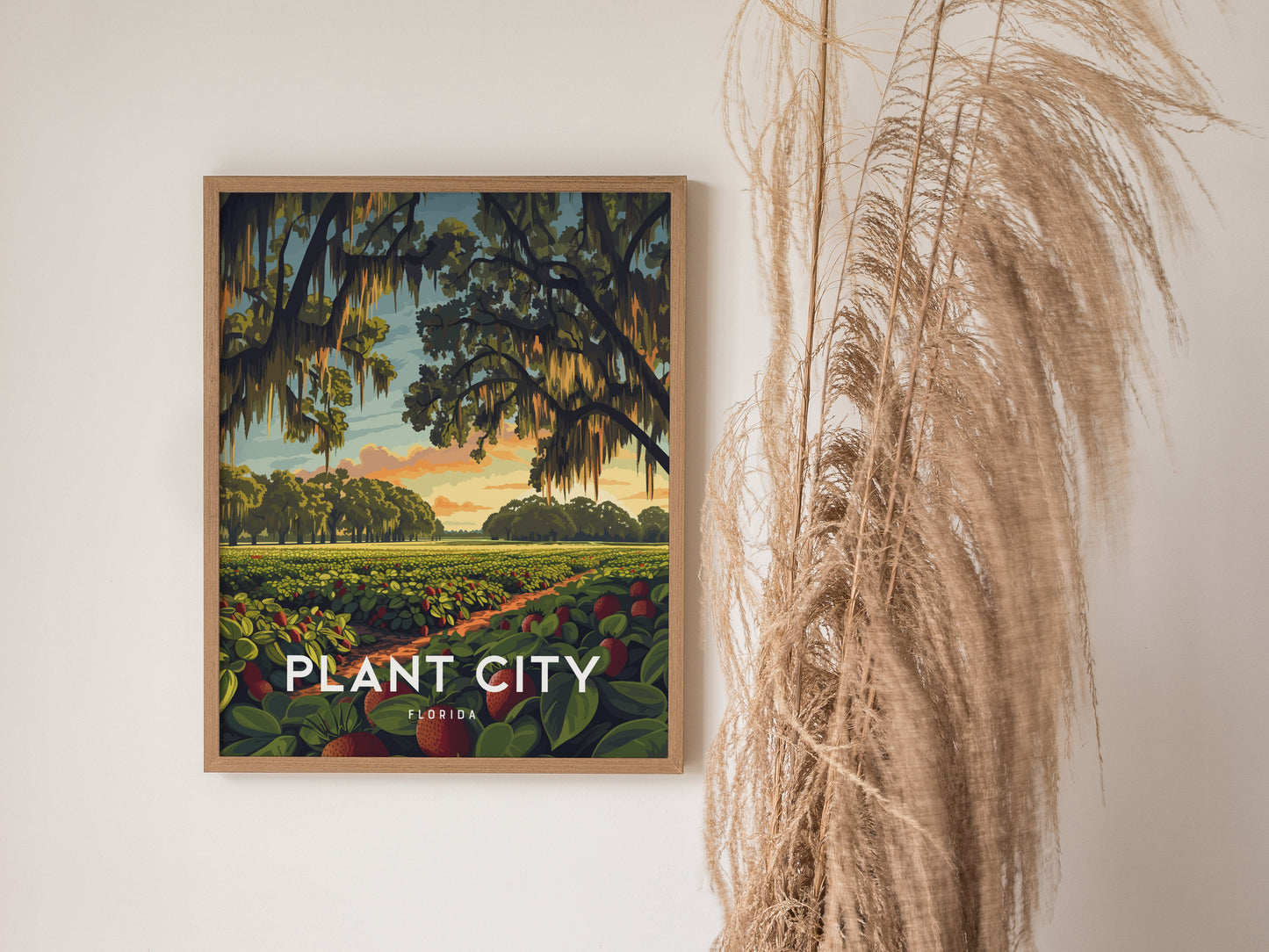 A framed poster of Plant City, Florida, depicting lush strawberry fields under a sunrise sky with oak trees draped in Spanish moss, hangs on a wall beside dried pampas grass.