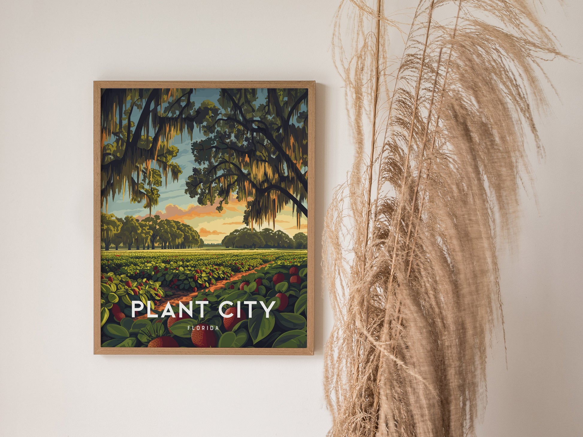 A framed poster of Plant City, Florida, depicting lush strawberry fields under a sunrise sky with oak trees draped in Spanish moss, hangs on a wall beside dried pampas grass.