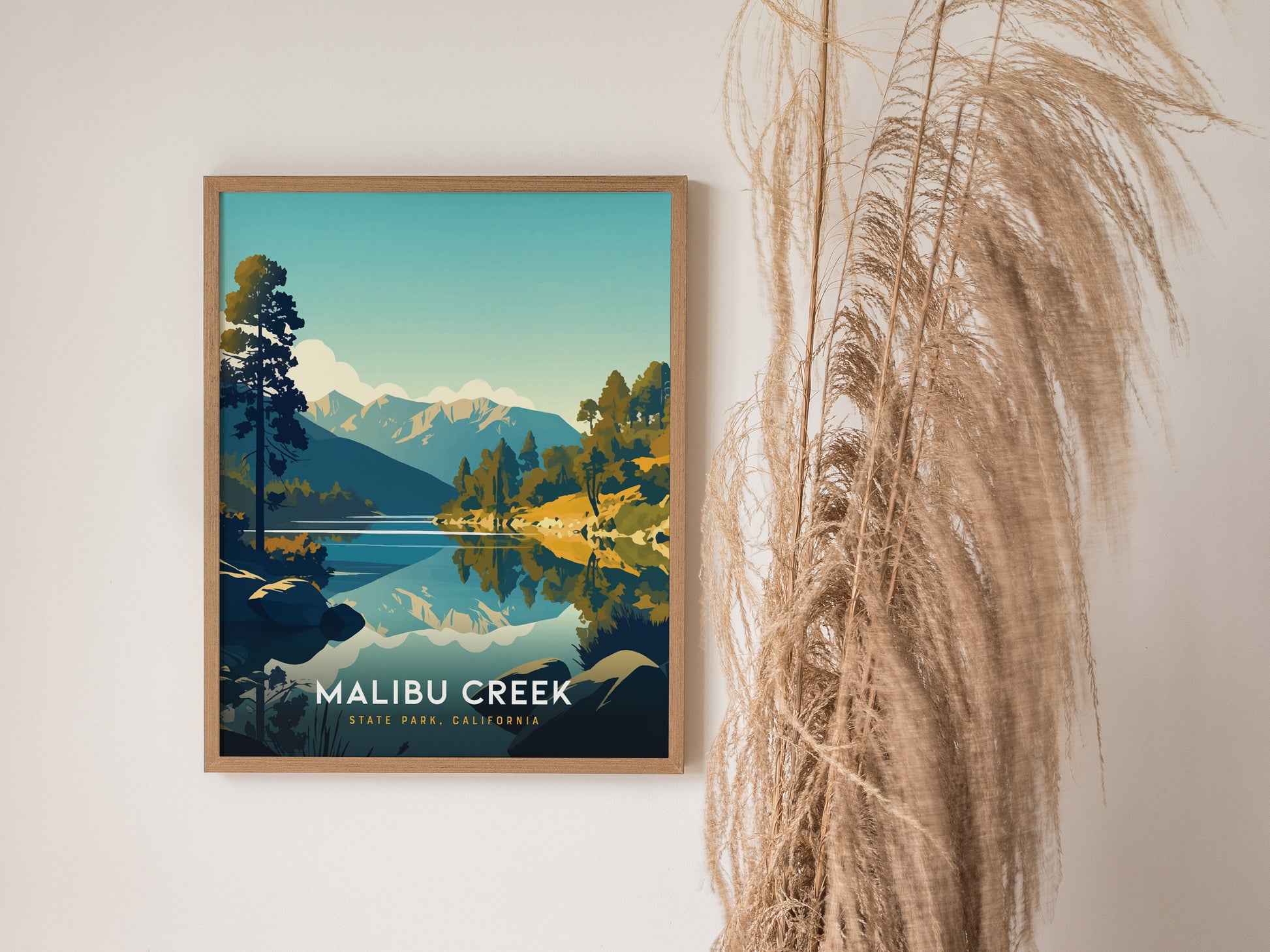 A poster of Malibu Creek State Park features illustrated mountains and a reflective creek, framed in wood. Its displayed on a wall beside tall, dried grasses.