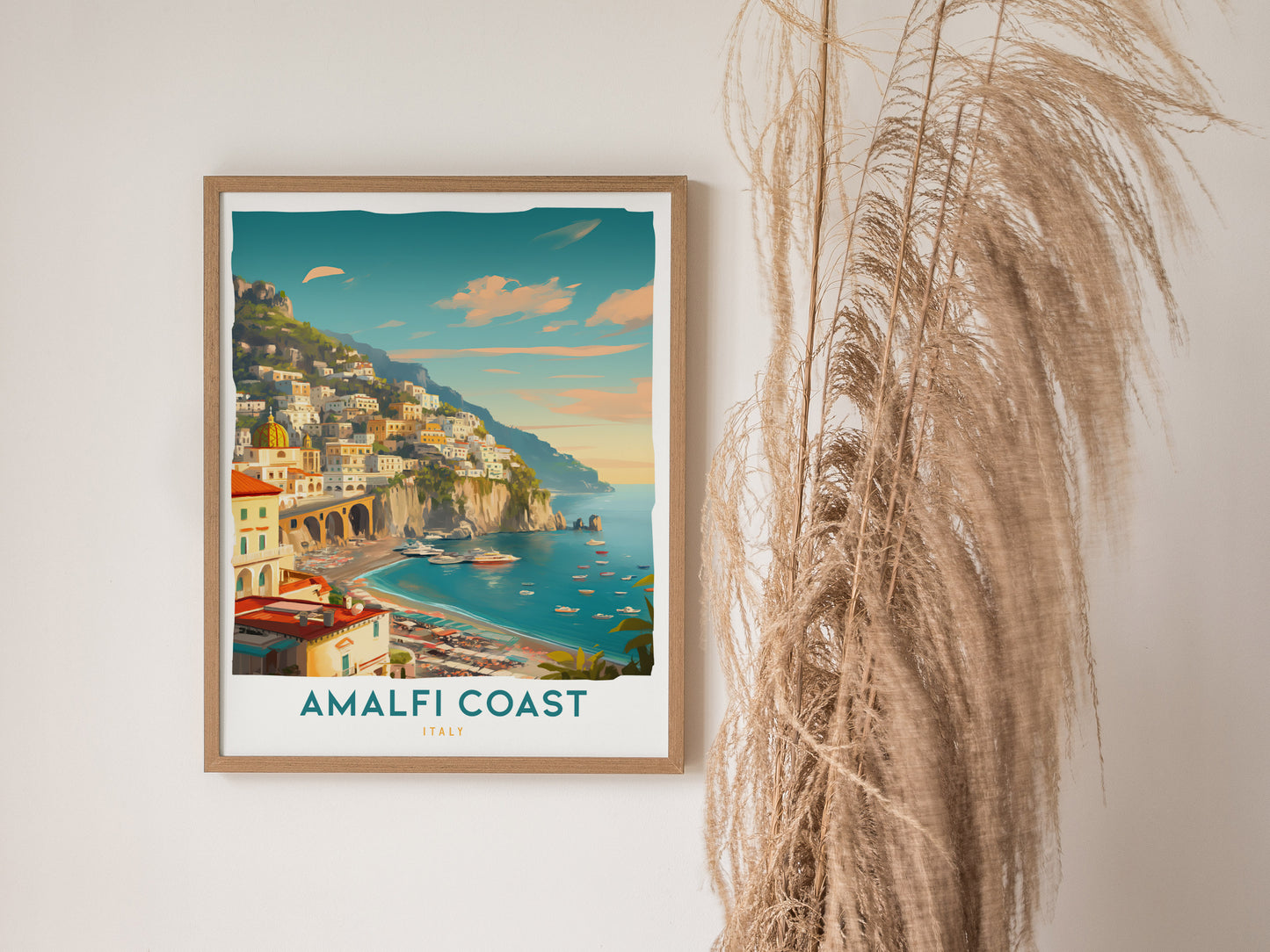 A framed Amalfi Coast travel print hangs on a white wall, depicting colorful seaside villages and boats. Tall dried grasses are visible beside the artwork.