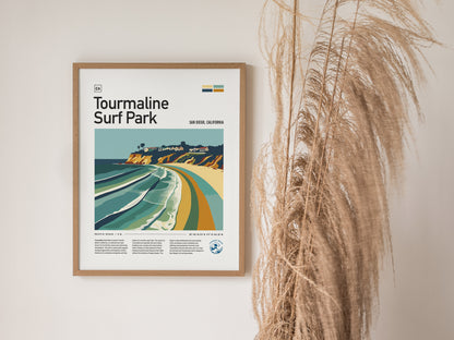 Framed poster of Tourmaline Surf Park featuring a stylized coastal scene hangs on a wall beside tall, dried pampas grass. The artwork showcases vibrant waves and cliffs.
