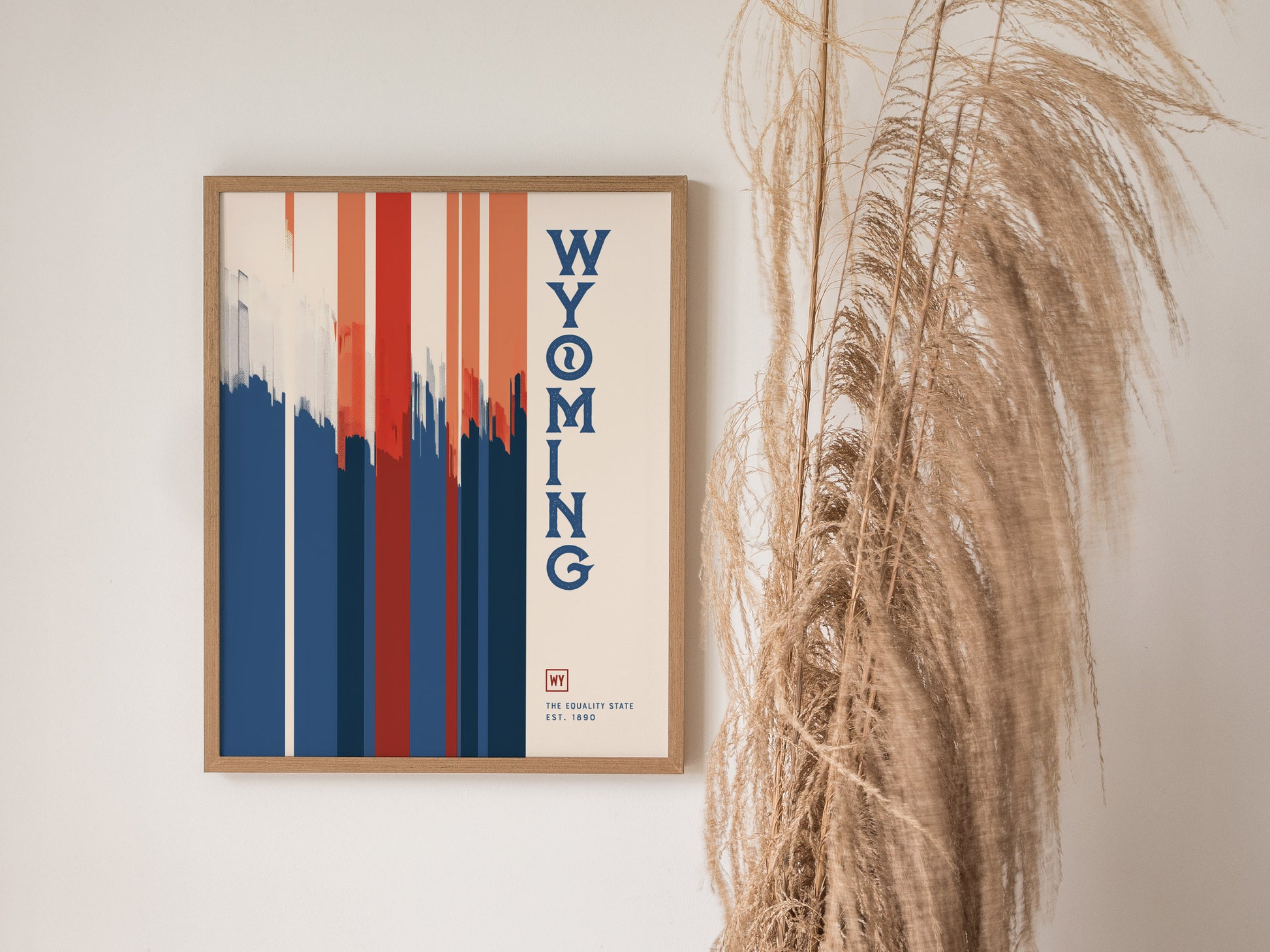 A framed Wyoming poster featuring bold red, white, and blue vertical stripes with the word WYOMING in blue text. Pampas grass decorates the right side of the image.