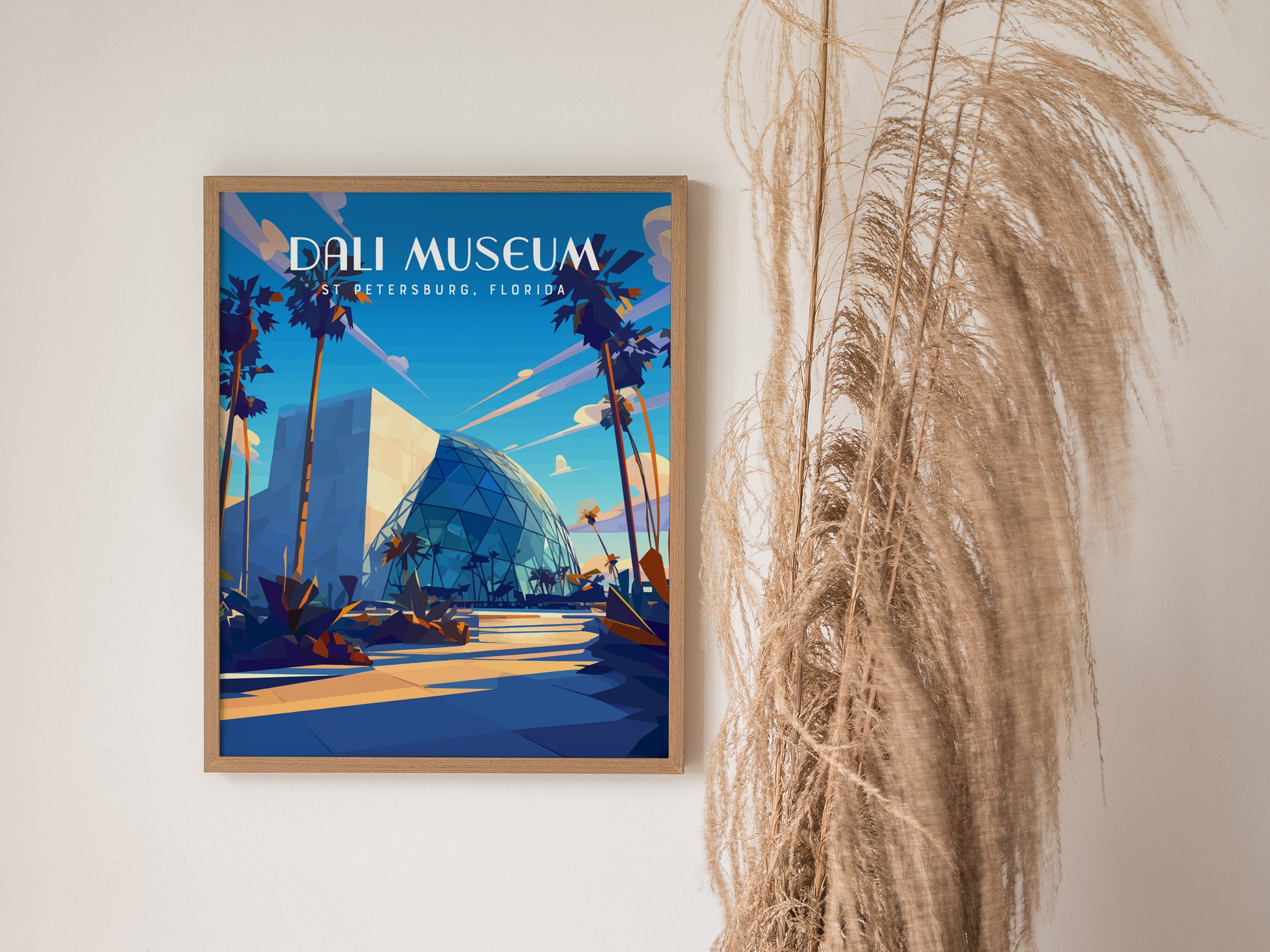 A framed poster of the Dali Museum in St. Petersburg, Florida, featuring modern architecture with a glass atrium, is displayed against a white wall beside decorative dried grasses.