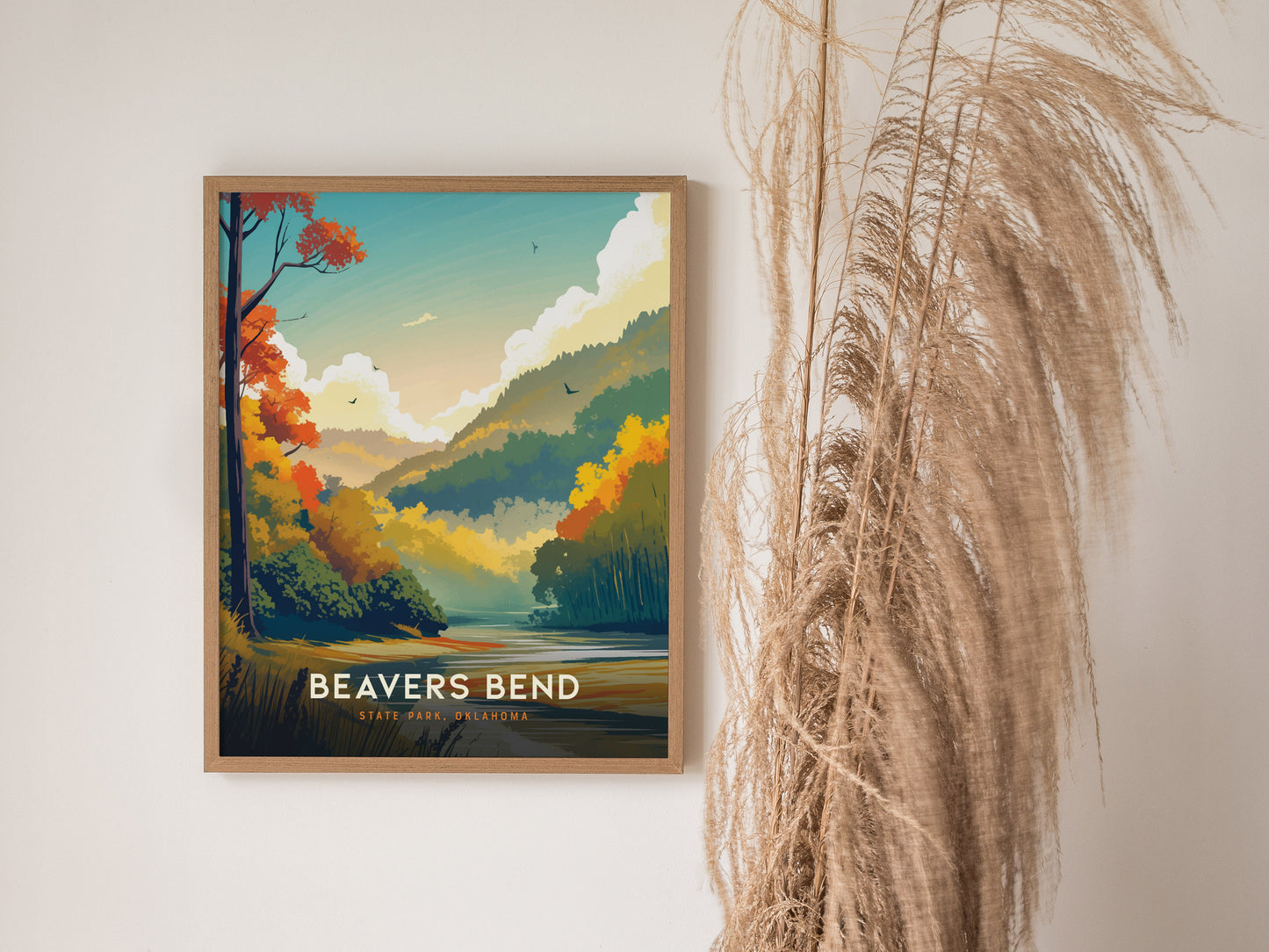 A framed poster of Beavers Bend State Park features vibrant autumn colors with trees, hills, and a serene river. It is displayed on a wall beside tall dried grasses.