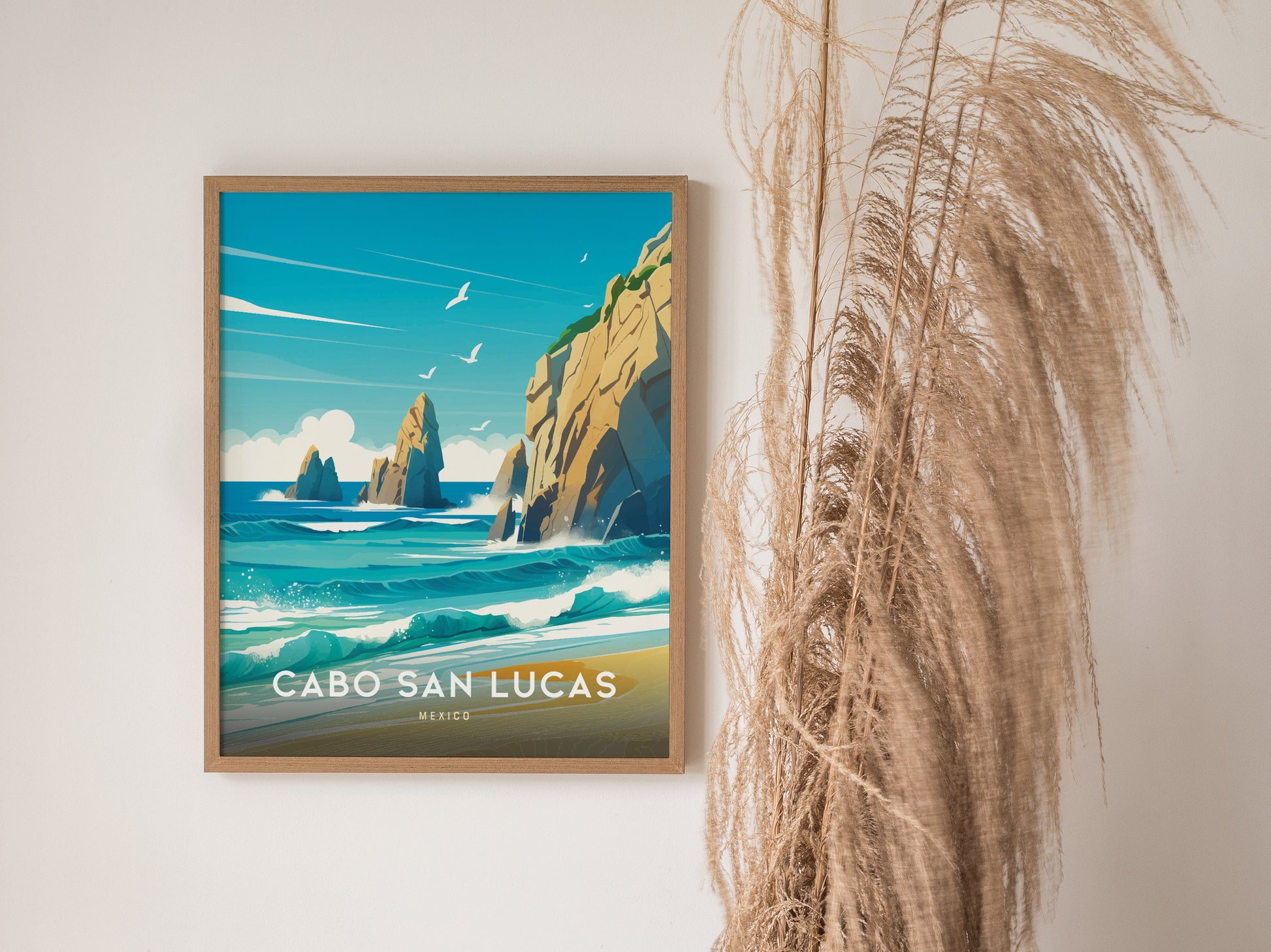 A framed Cabo San Lucas poster hangs on a white wall, depicting turquoise waves and rocky cliffs under a blue sky. Pampas grass decorates the space beside it.