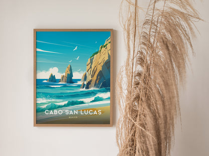 A framed Cabo San Lucas poster hangs on a white wall, depicting turquoise waves and rocky cliffs under a blue sky. Pampas grass decorates the space beside it.