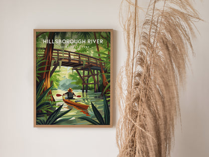 A framed poster of Hillsborough River State Park features a kayaker beneath a wooden bridge surrounded by lush greenery. It hangs on a wall next to tall, dried grass.