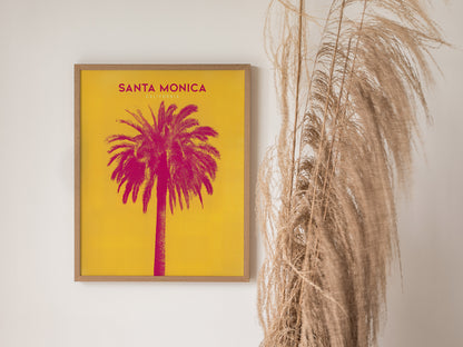 A framed poster featuring a vibrant pink palm tree against a yellow background with Santa Monica California text. The poster is displayed next to tall, dried pampas grass.