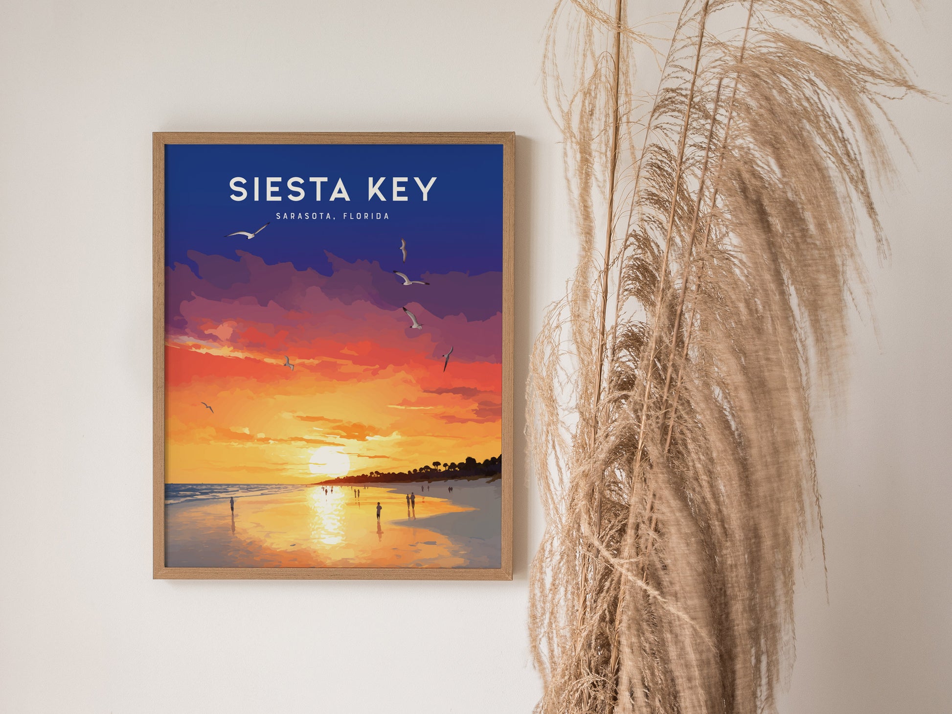 A framed poster of a vibrant sunset over a beach, with silhouettes of people and birds, hangs on a wall beside tall dried grasses.