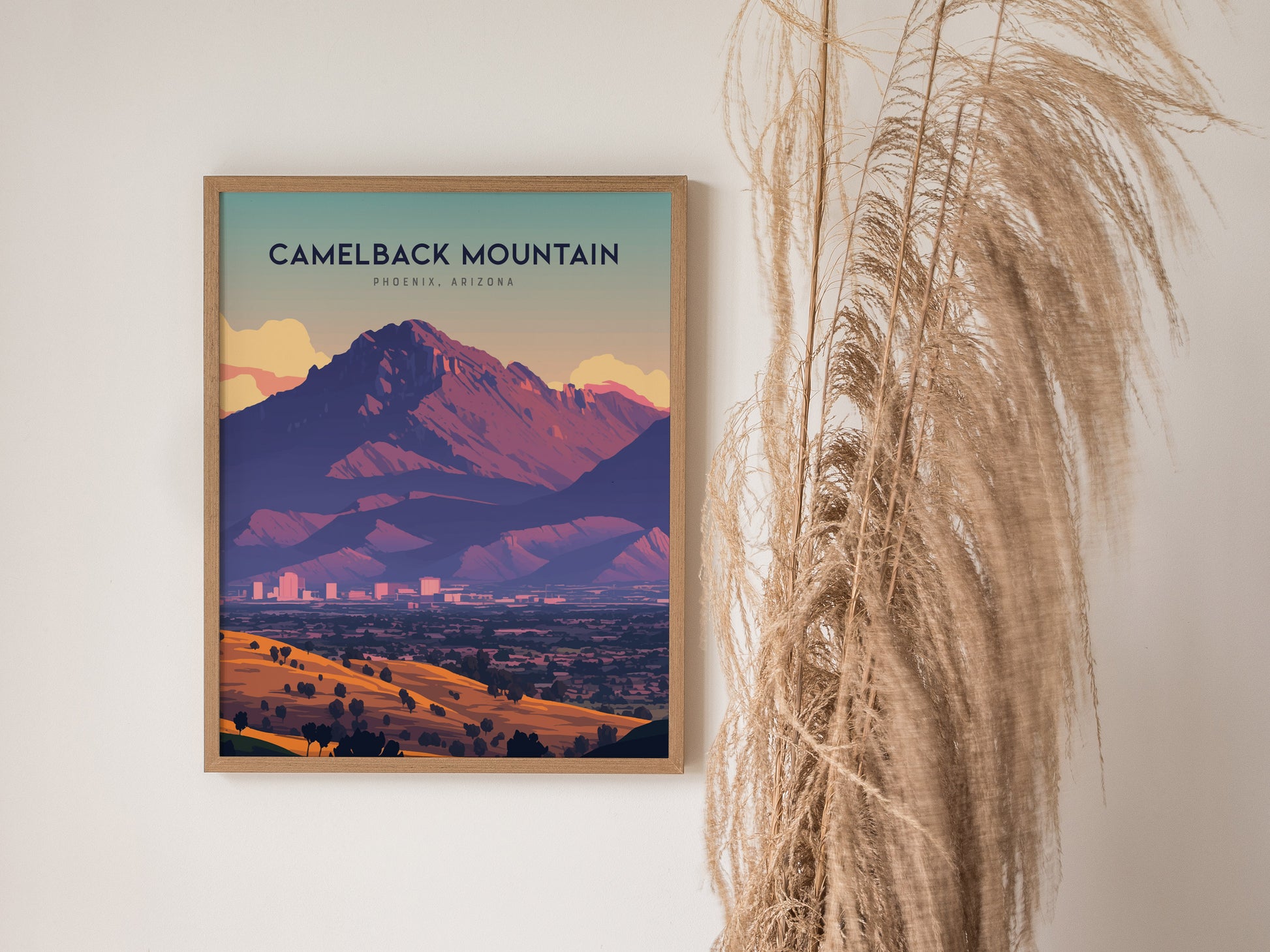 A framed poster of Camelback Mountain in Phoenix, Arizona, hangs on a white wall next to tall, dried grasses, showcasing vibrant desert colors and a dramatic silhouette.