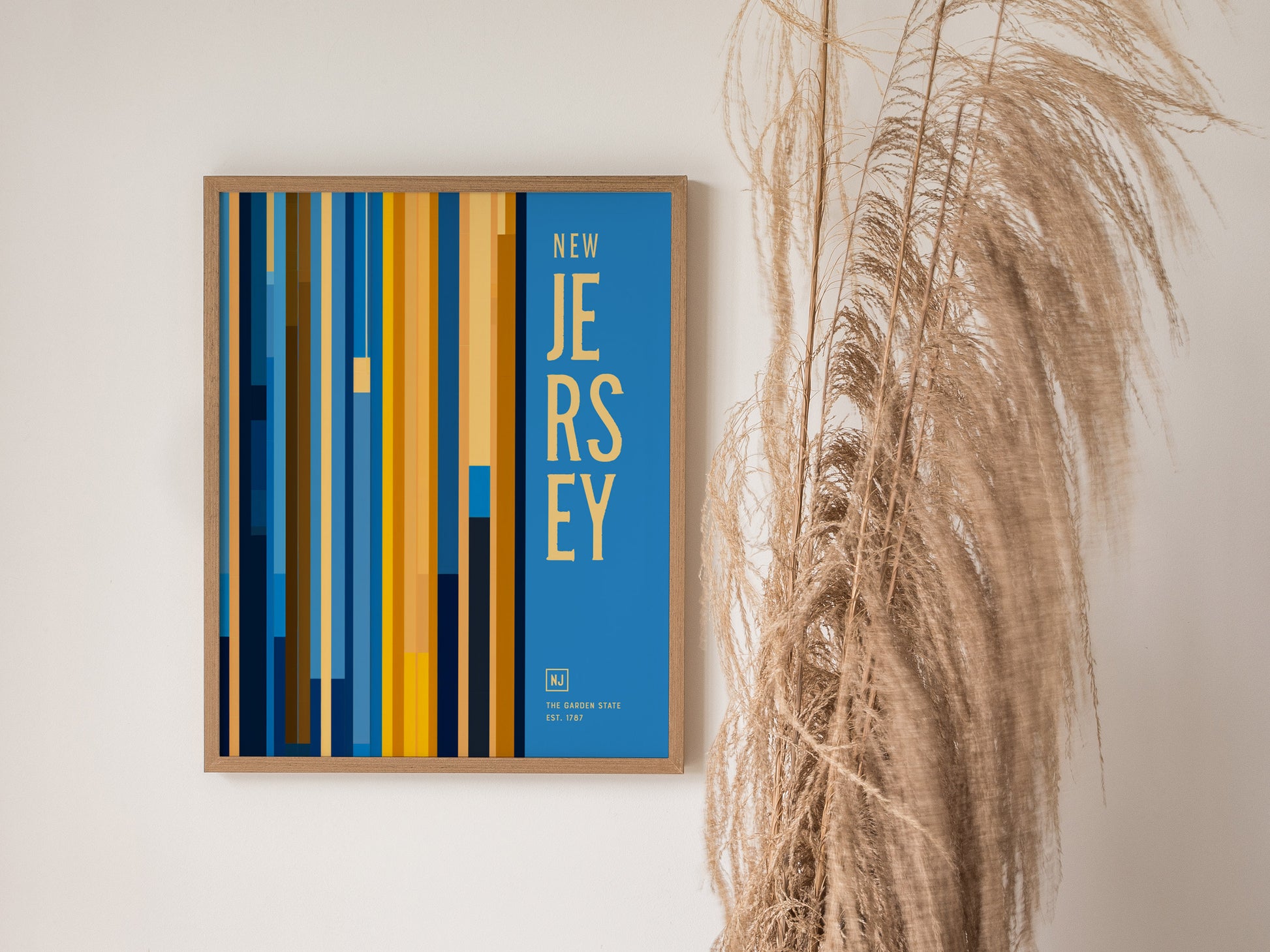 A framed New Jersey poster with vertical blue and yellow stripes and bold text, hanging on a wall next to tall, dried pampas grass.