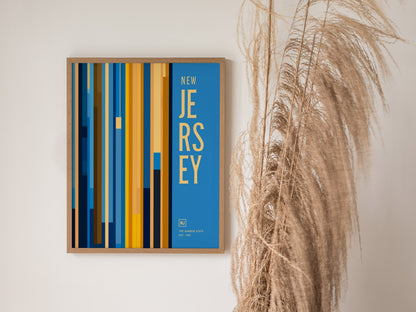 A framed New Jersey poster with vertical blue and yellow stripes and bold text, hanging on a wall next to tall, dried pampas grass.