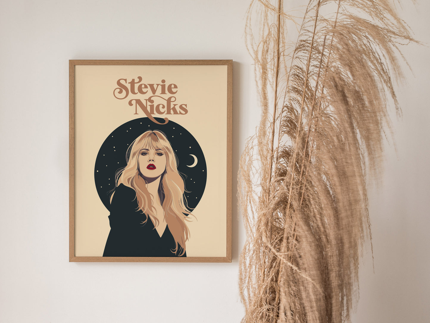 A framed poster of Stevie Nicks features her portrait with a celestial background, including stars and a crescent moon, next to decorative dried pampas grass.
