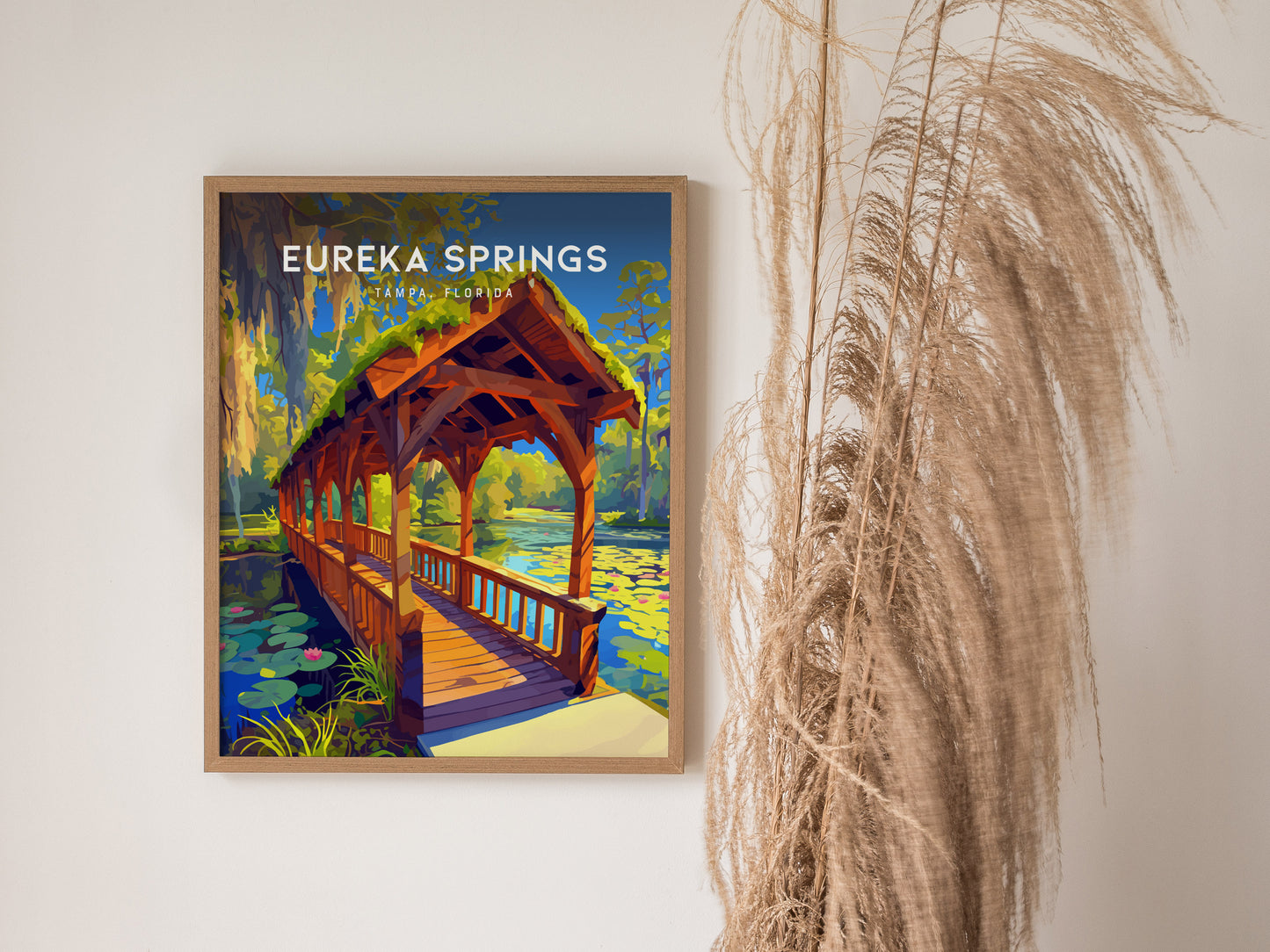 A framed Eureka Springs poster depicting a wooden bridge over a stream with water lilies and Spanish moss. Its displayed on a wall next to dried pampas grass.