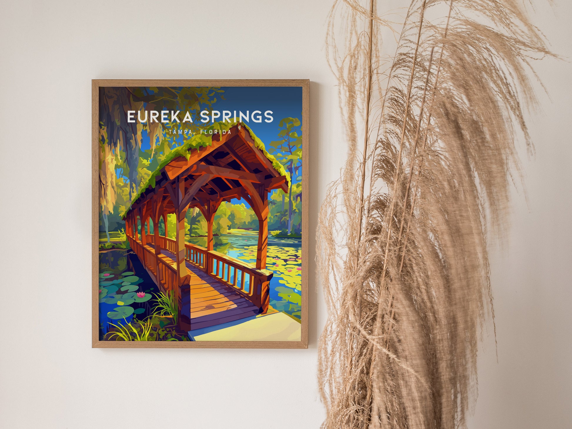 A framed Eureka Springs poster depicting a wooden bridge over a stream with water lilies and Spanish moss. Its displayed on a wall next to dried pampas grass.