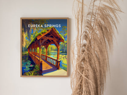 A framed Eureka Springs poster depicting a wooden bridge over a stream with water lilies and Spanish moss. Its displayed on a wall next to dried pampas grass.